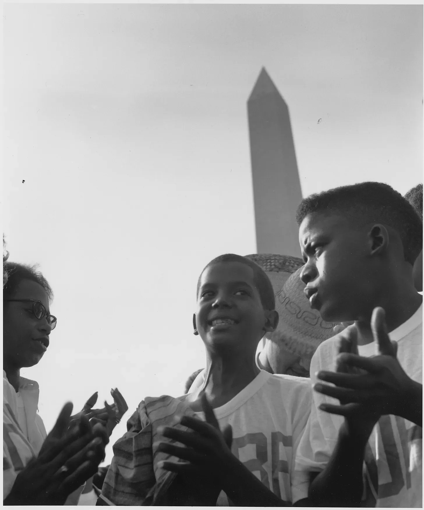 three black children stand close together, perhaps playing a hand-clapping game. the tall pointed shape of the washington monument is visible in the background.