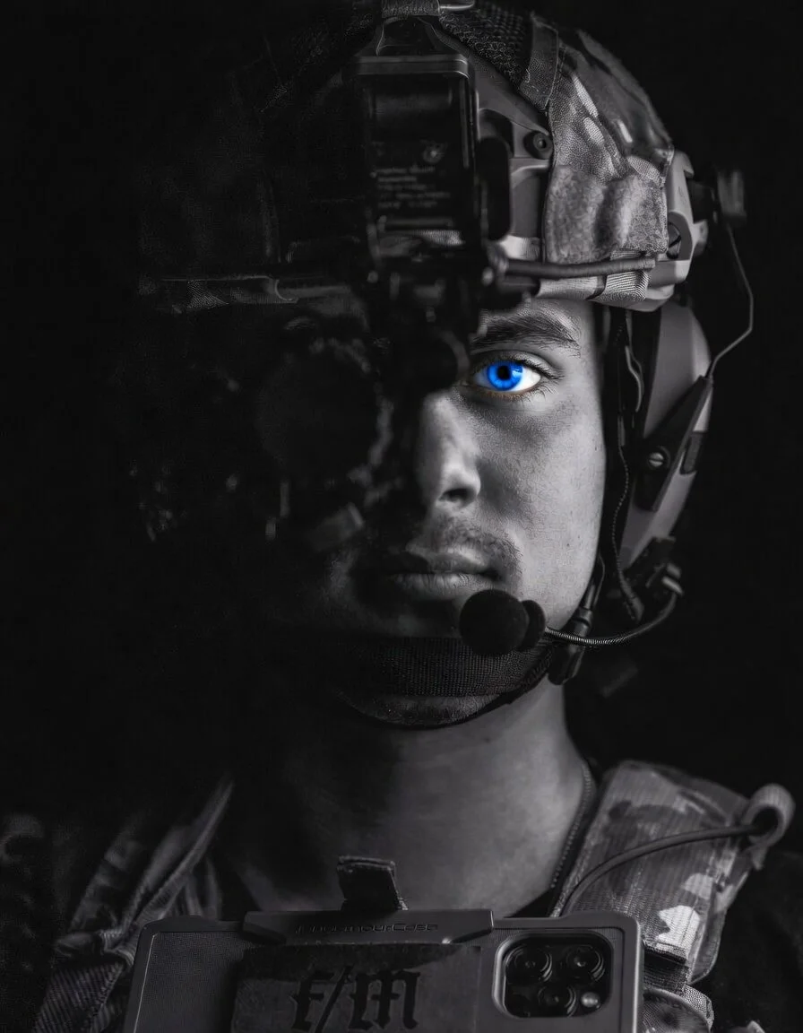 A soldier in military gear wearing a camo uniform and headgear with a headset, holding a smartphone in front of him, with his right eye visible, glowing blue. The photo is in black and white except for his eye.