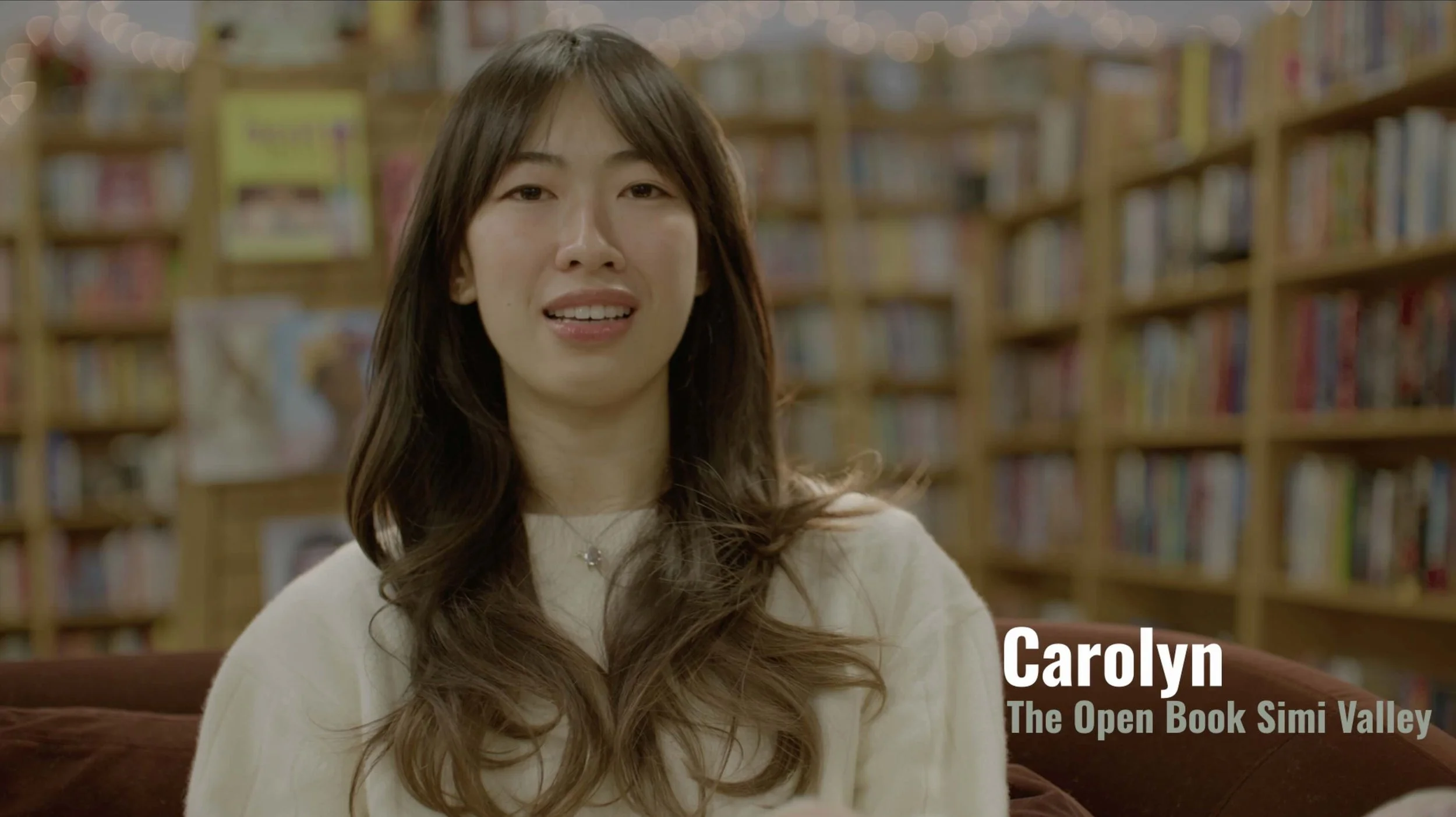 A woman with long brown hair wearing a white top and a necklace, sitting in a library with wooden bookshelves full of books in the background.