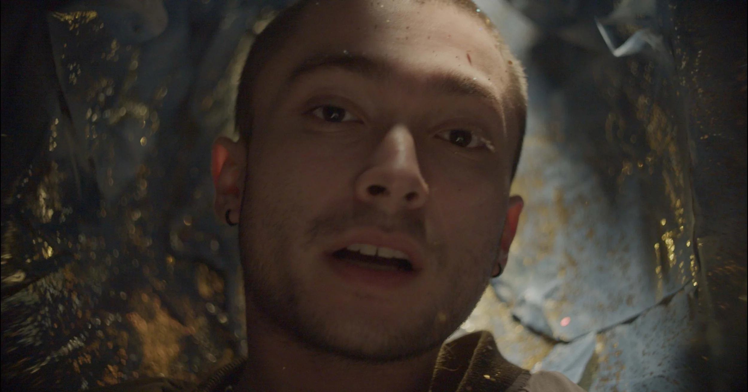 A musical artist lies inside a reflective, metallic cocoon with gold spots, looking directly at the camera while performing a song.