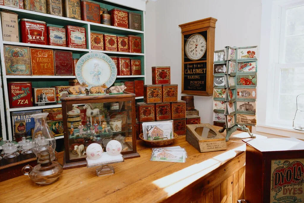 A vintage shop display with shelf filled with antique tea boxes, a glass cabinet with small figurines, and a wooden table with miscellaneous collectibles and artwork. There's a wall clock and a postcard rack on the side.