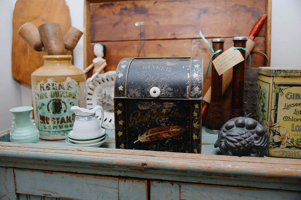 Vintage kitchen counter with various old utensils and containers, including a black miniature stove, ceramic jars, a small pitcher, and a decorative pig figurine.
