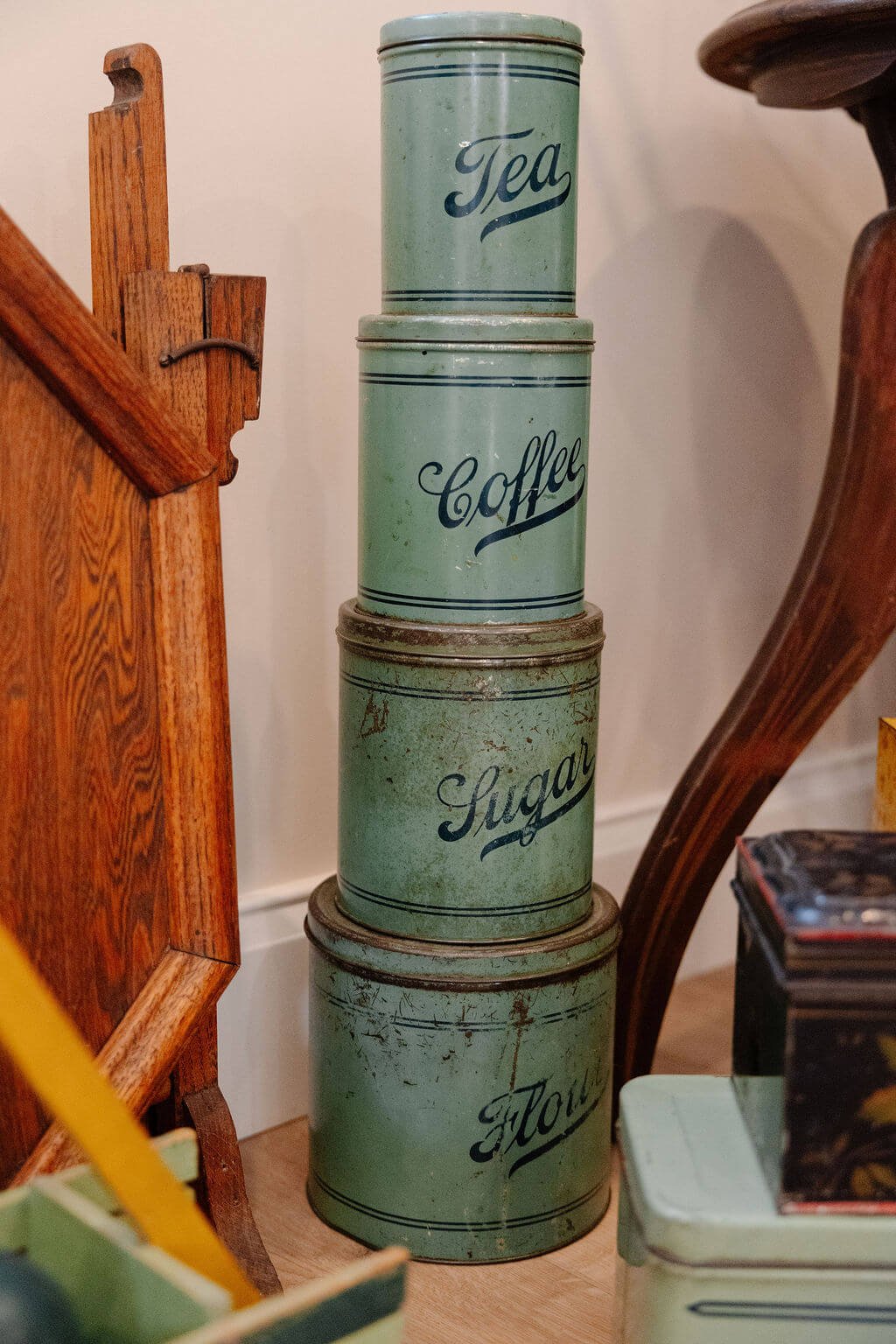 Four vintage cylindrical tins stacked on top of each other with labels reading 'Tea,' 'Coffee,' 'Sugar,' and 'Flour' from top to bottom, placed against a white wall.