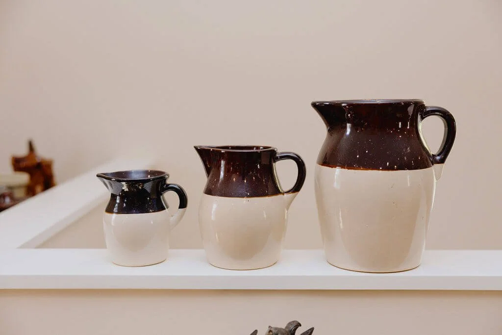 Three ceramic pitchers with a beige lower part and dark brown upper part on a white shelf.