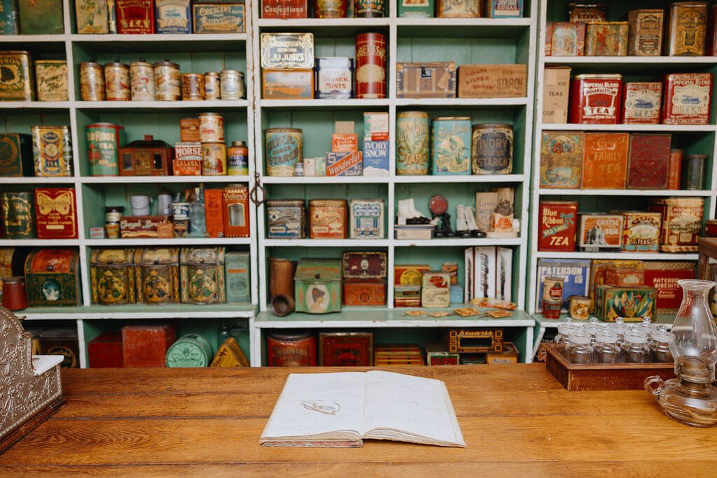 Vintage tea tins and boxes on shelves behind a wooden counter, with an open notebook and glass oil lamp on the counter.