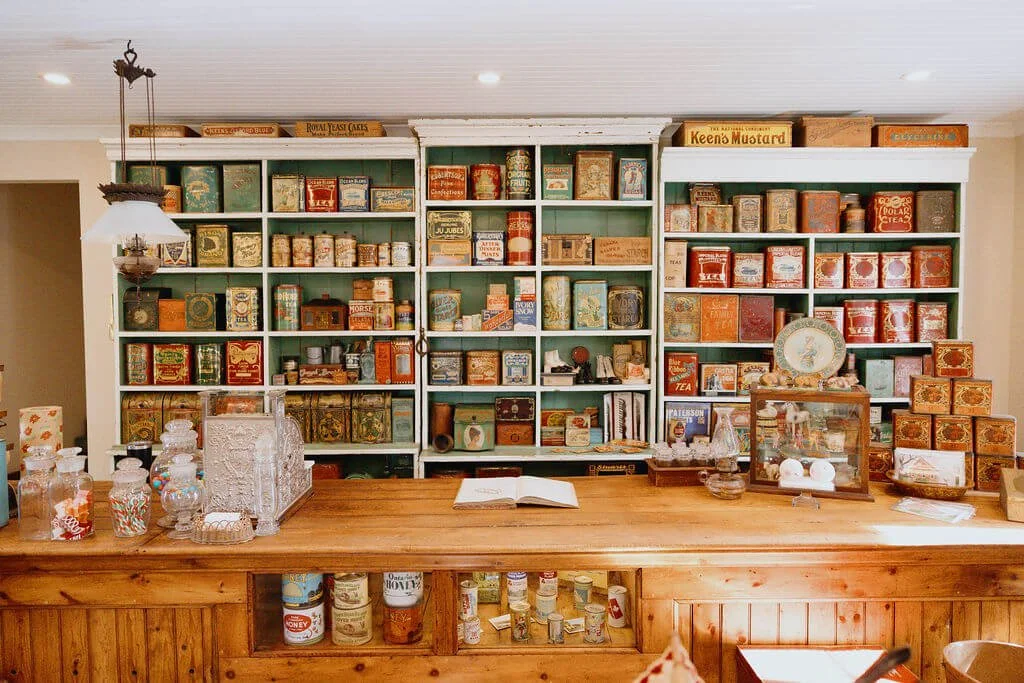 Vintage wooden counter with glass jars and decorative items, behind a large green shelving unit filled with vintage tins and cans, in a cozy shop or kitchen setting.