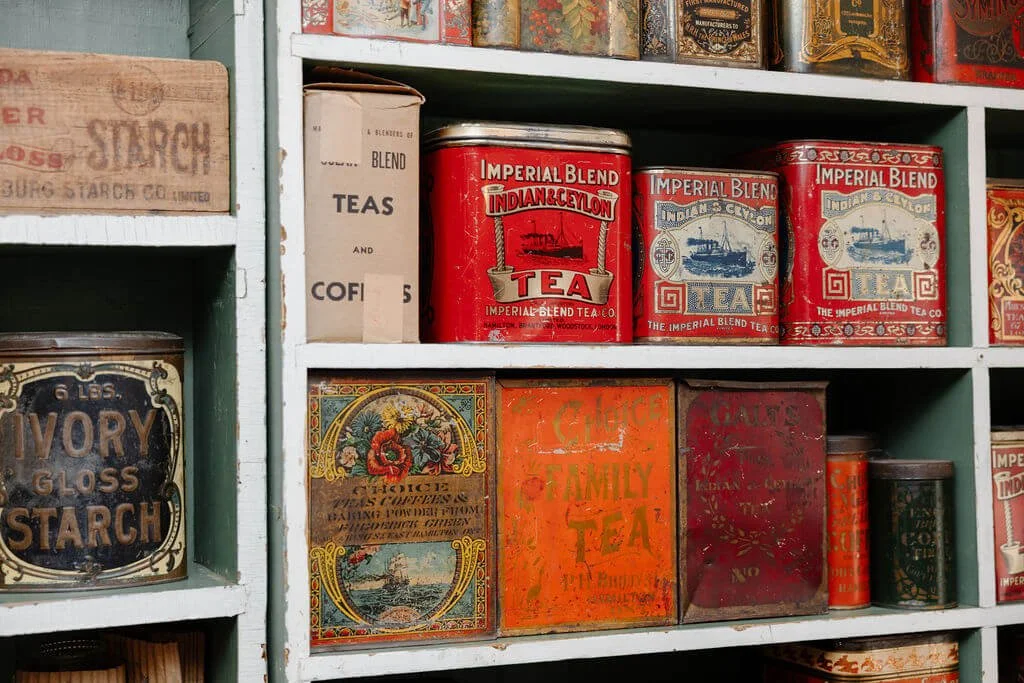 A shelf filled with vintage tea and starch tins, including Imperial Blend India & Ceylon tea tins, Ivory Gloss Starch, and other antique packaging.