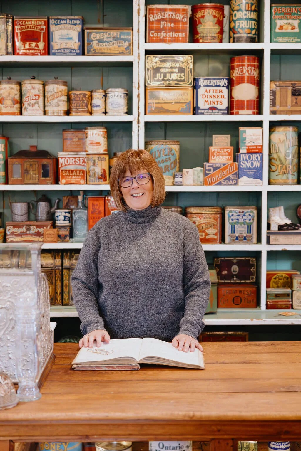A smiling woman with glasses and shoulder-length brown hair in a gray turtleneck sweater, standing behind a wooden counter with an open book, in front of a colorful vintage shop filled with tins and boxes on shelves.