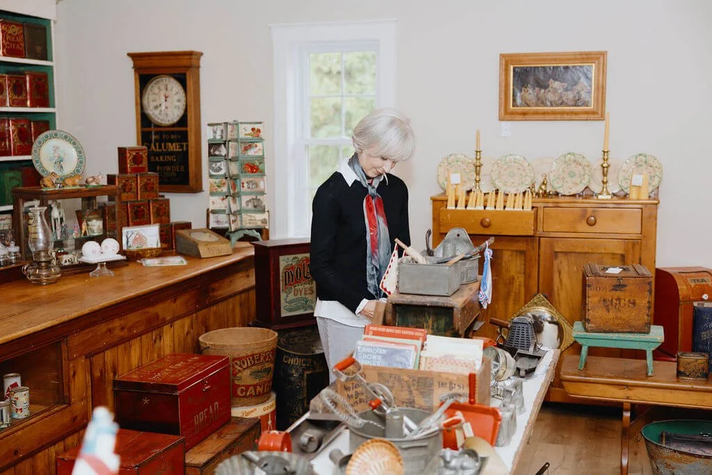 An elderly woman with white hair wearing a dark sweater over a light shirt, browsing items at an antique shop. The shop has wooden furniture and shelves filled with vintage collectibles and decorative plates.