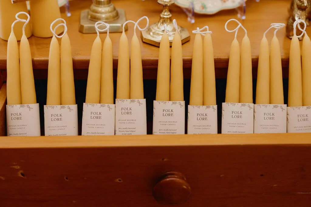 Several cream-colored taper candles with tags reading 'Folk Lore' on a wooden surface, likely for sale or display.