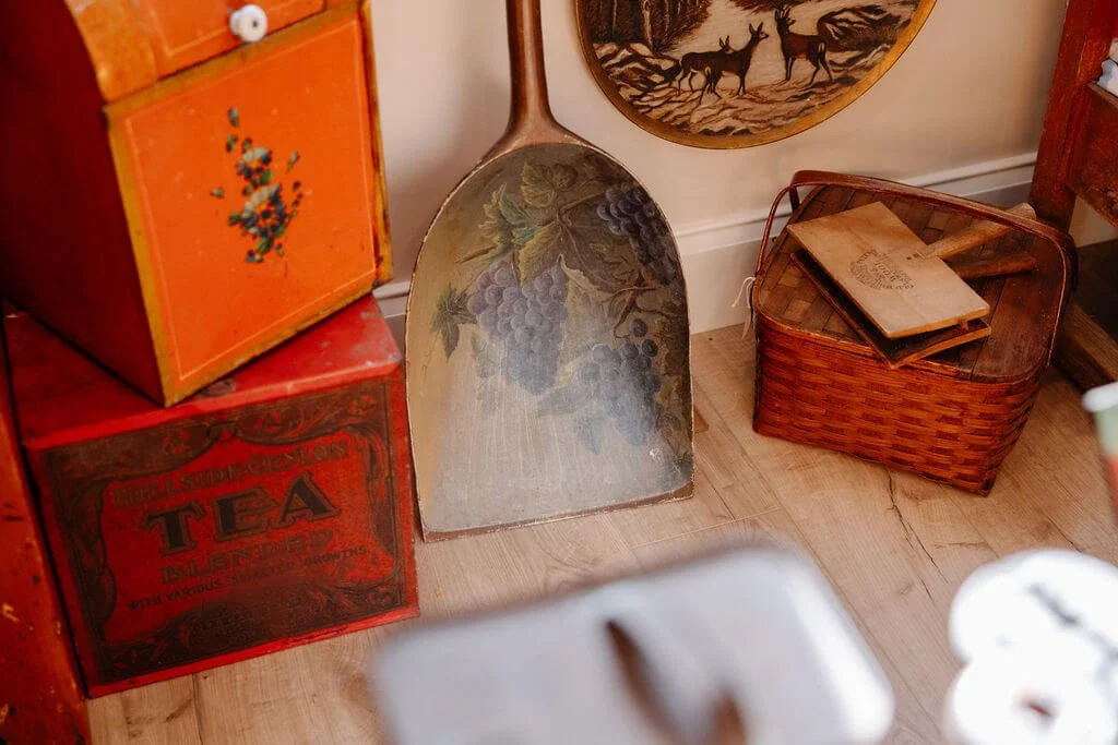Vintage kitchen decor with a large spoon featuring a grapevine design, wooden boxes, and a woven basket with old books on top, all on a wooden floor.
