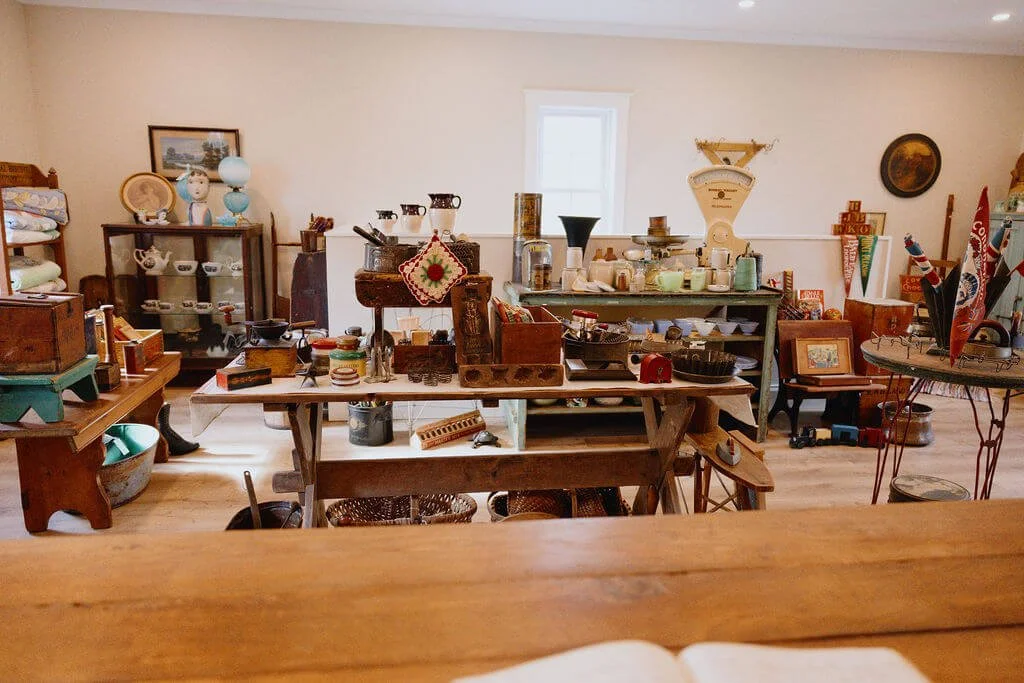 A room filled with vintage and antique items such as pottery, scales, boxes, and decorative objects, arranged on tables and shelves in a cozy setting with a window in the background.