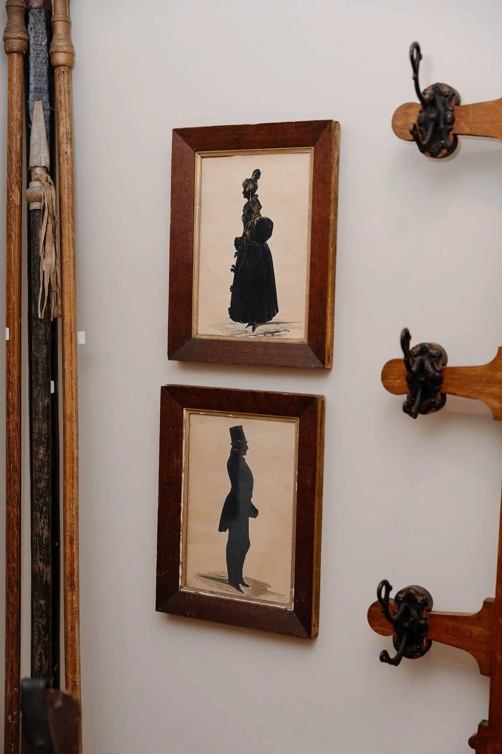 Two vintage sepia-toned silhouette portraits of people in old-fashioned clothing, framed and hung vertically on a wall. To the left of the frames, part of a wooden object and some antique tools are visible. To the right, part of a wooden coat rack wi