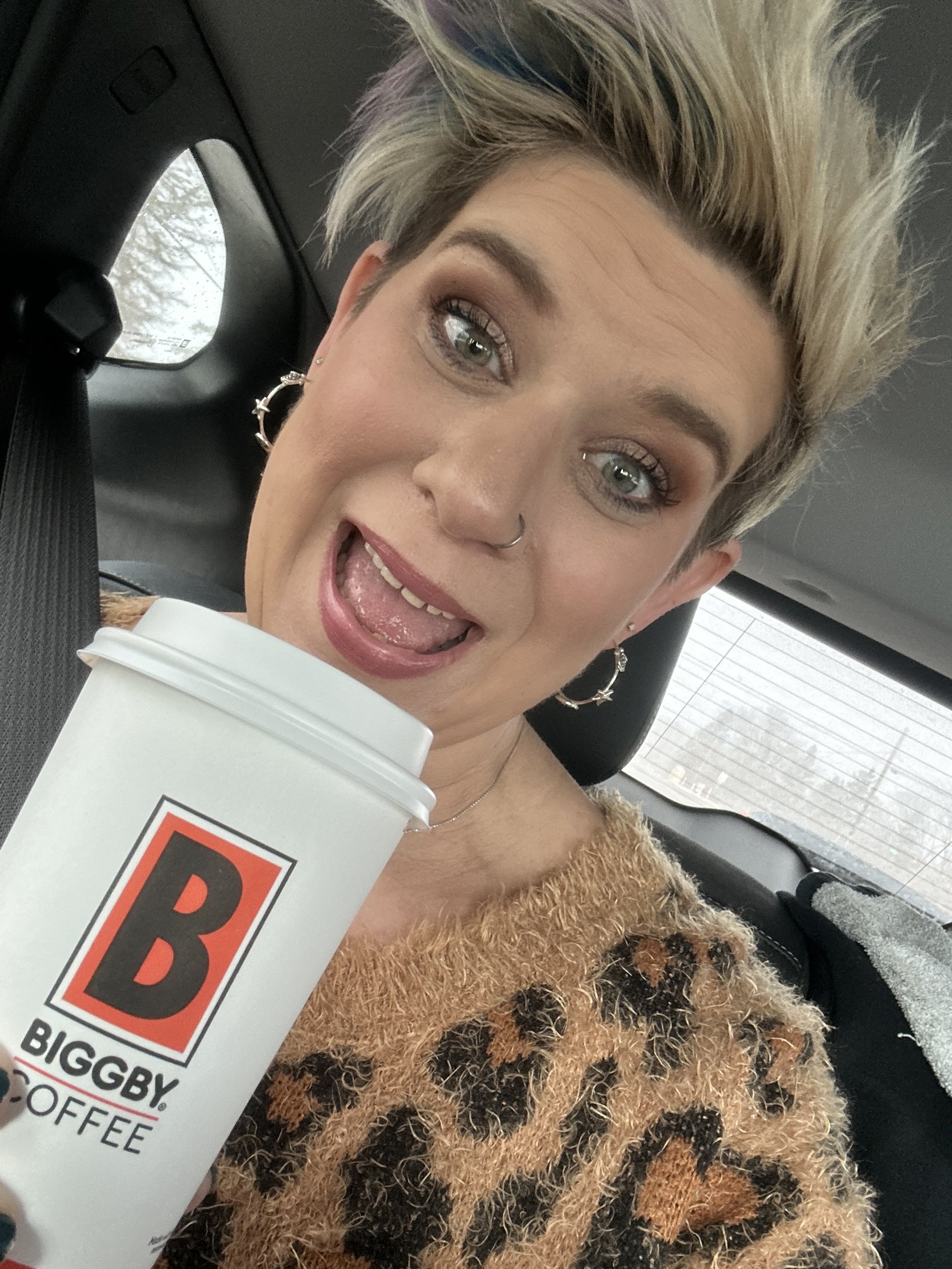 A woman with short blonde hair styled in a spiky fashion, wearing hoop earrings and a leopard print fuzzy sweater, holding a large white cup of Biggby Coffee, sticking her tongue out playfully in a car.