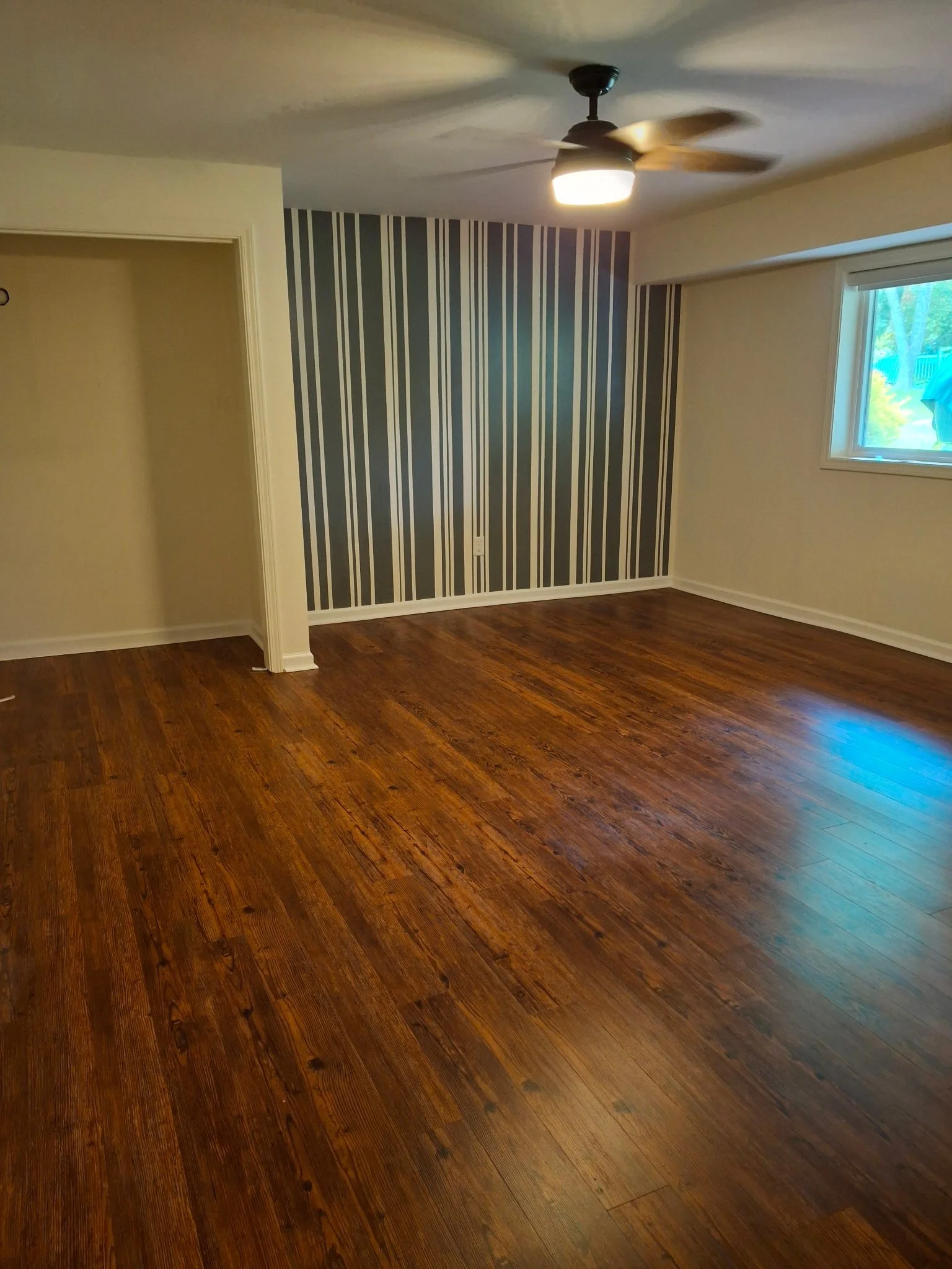 Empty room with hardwood floors, a ceiling fan with a light, a window on the right, and a wall with dark vertical striped wallpaper.