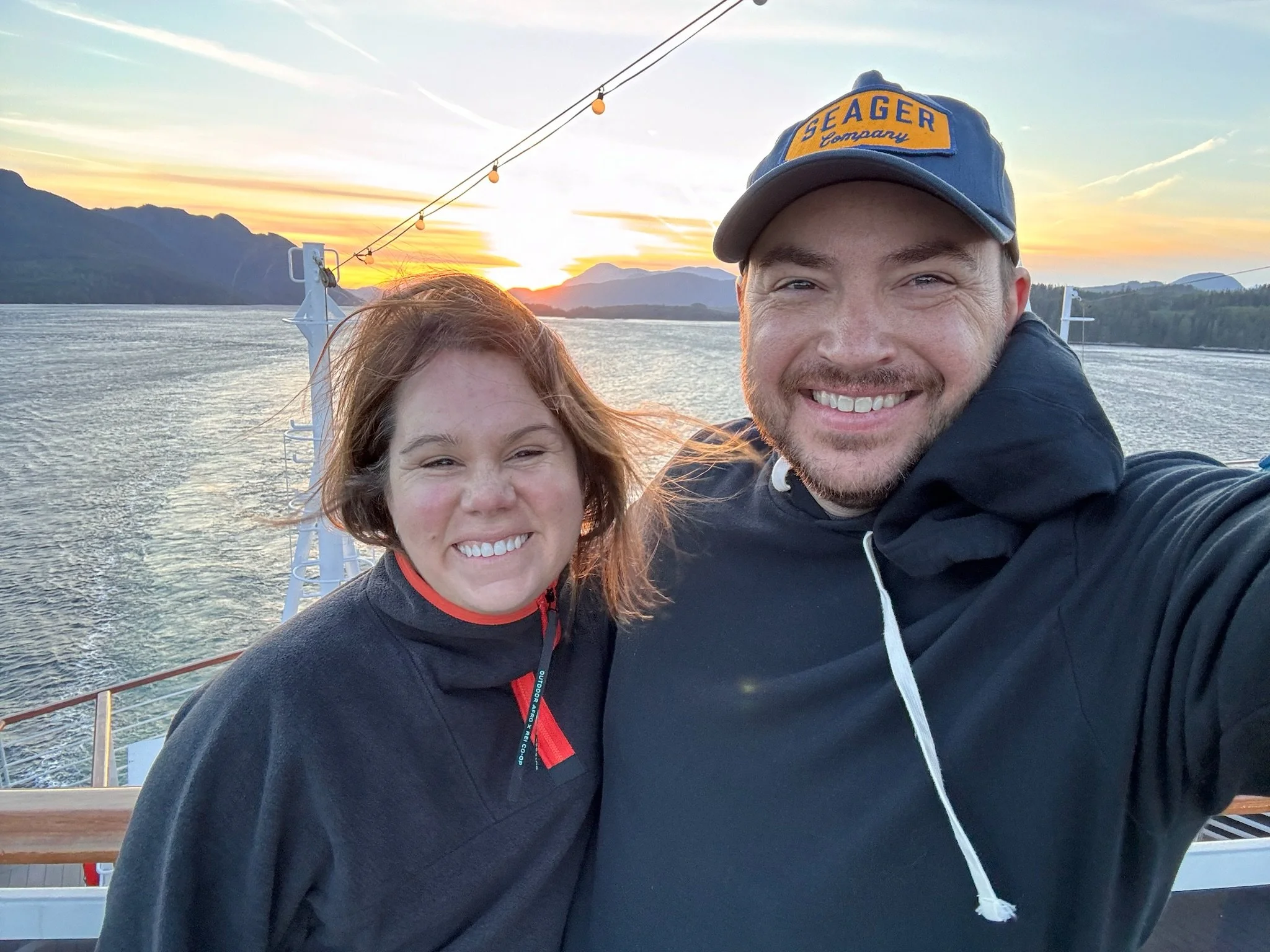 A smiling man and woman taking a selfie on a boat during sunset with mountains and water in the background.