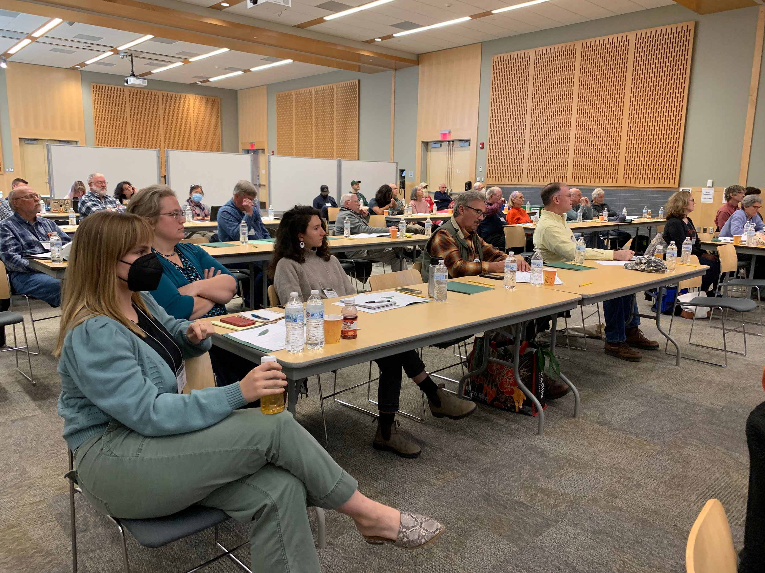 People attending a conference or workshop in a large room with beige walls, seated at tables with bottled water and notebooks.