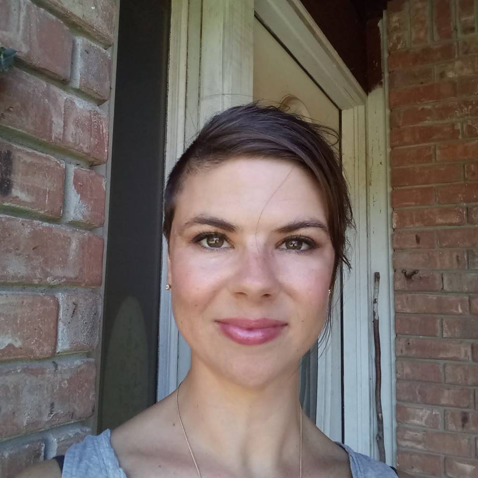 Close-up of a woman with short, dark hair smiling, standing outside near a brick wall and door.