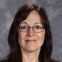 A woman with shoulder-length dark brown hair, wearing glasses, smiling in front of a gray background.