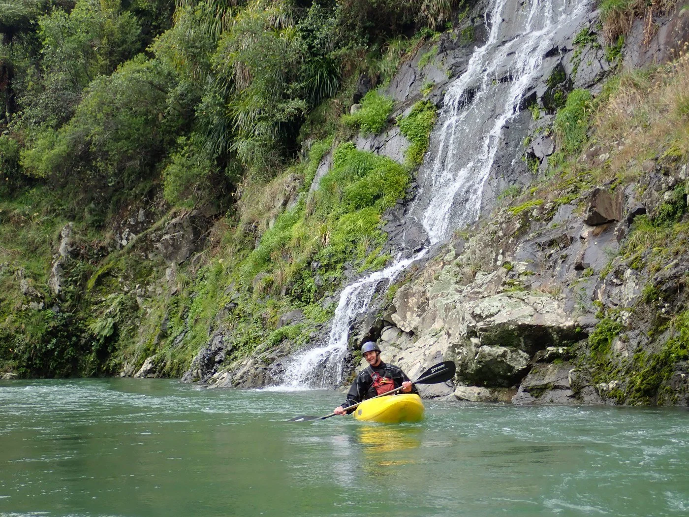 A person kayaking on a river with a waterfall and green foliage in the background.