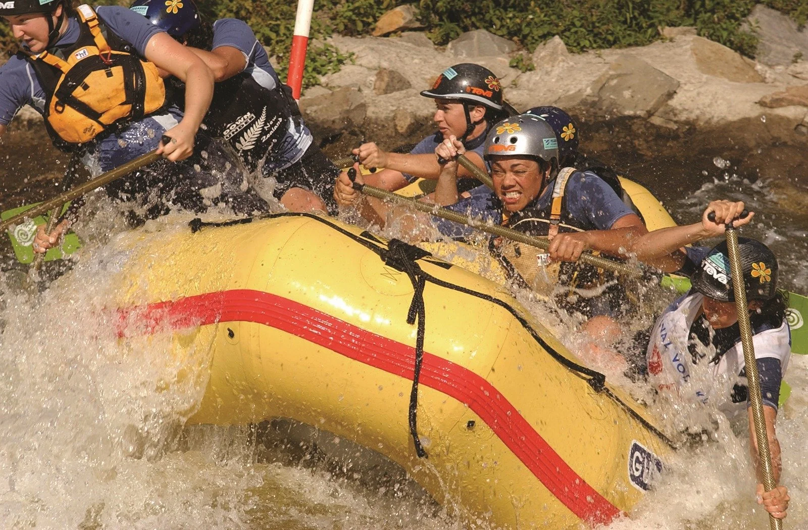 Group of people white water rafting in a yellow inflatable raft, wearing helmets and life jackets, paddling through rapid waters with rocky riverbank in background.