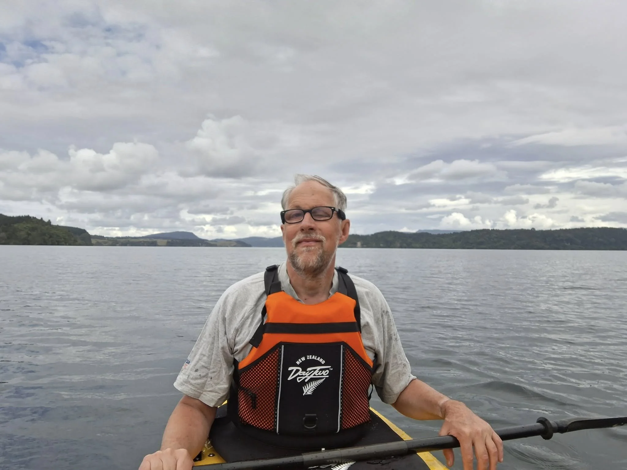A man with glasses and gray hair is kayaking on a lake under a cloudy sky, wearing a life jacket with "New Zealand" written on it.