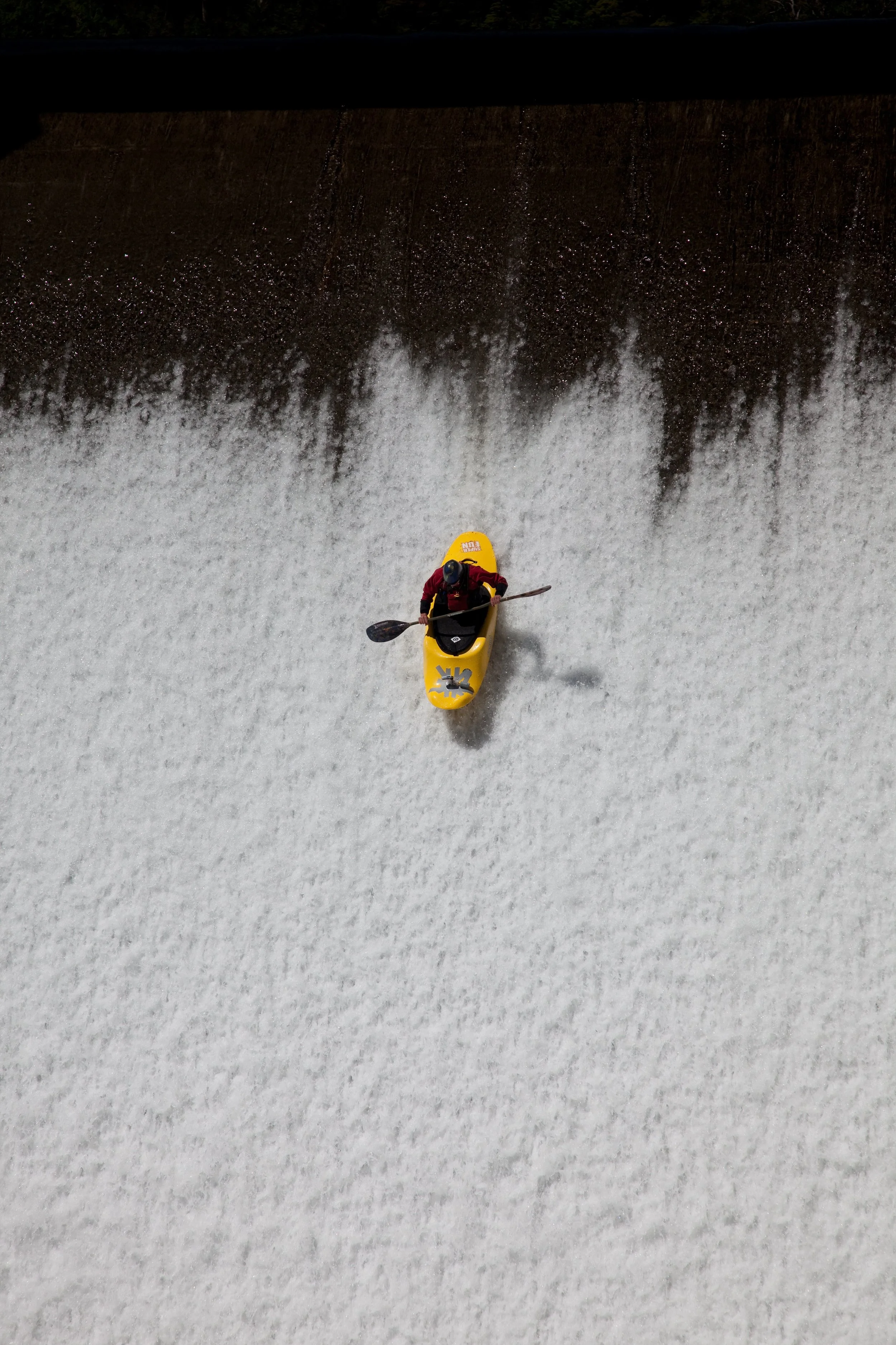 A person in a red jacket and helmet paddling a yellow kayak down a waterfall or rapid.