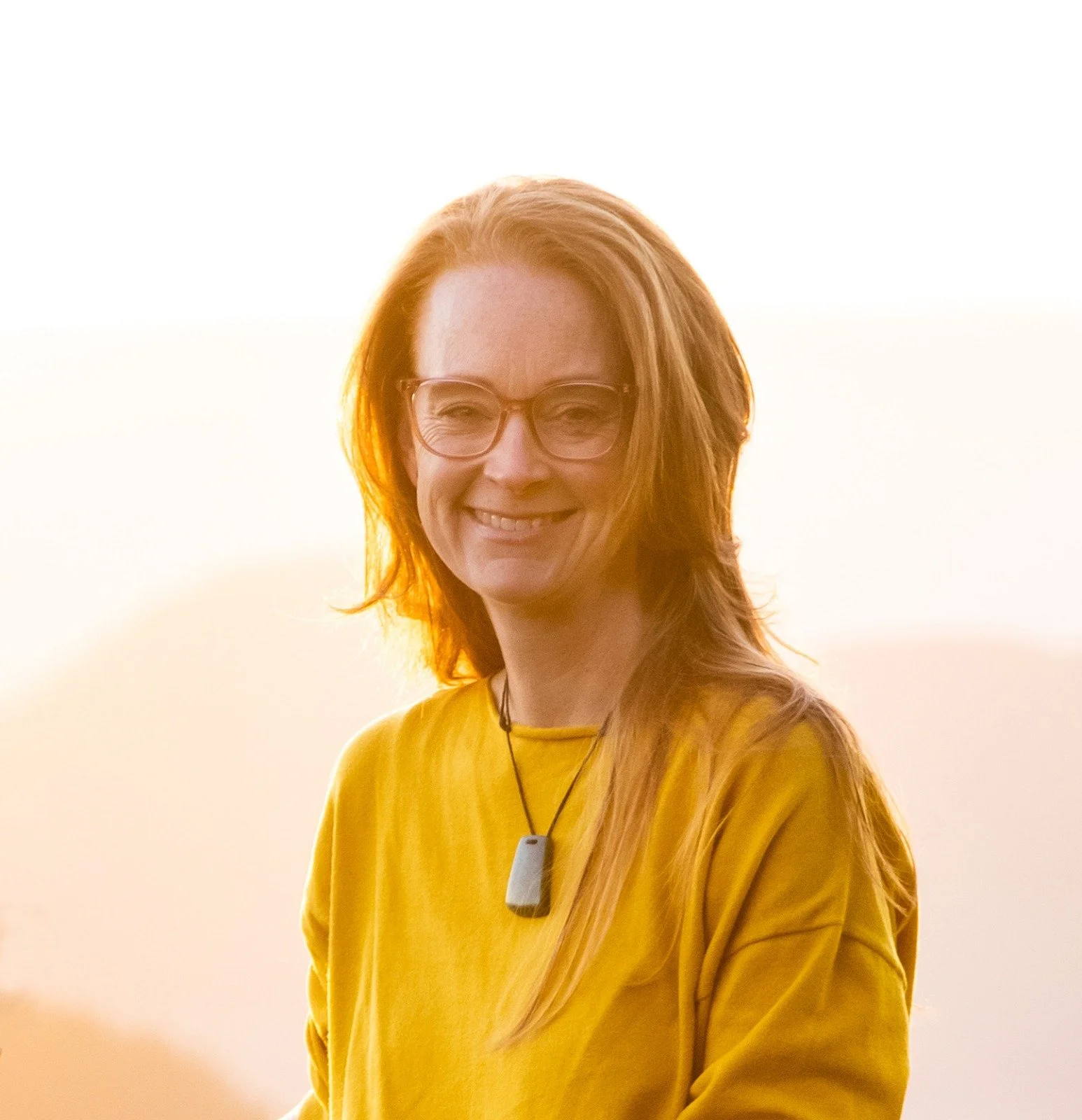 A woman with red hair and glasses smiling at sunset, wearing a yellow shirt and a pendant necklace.