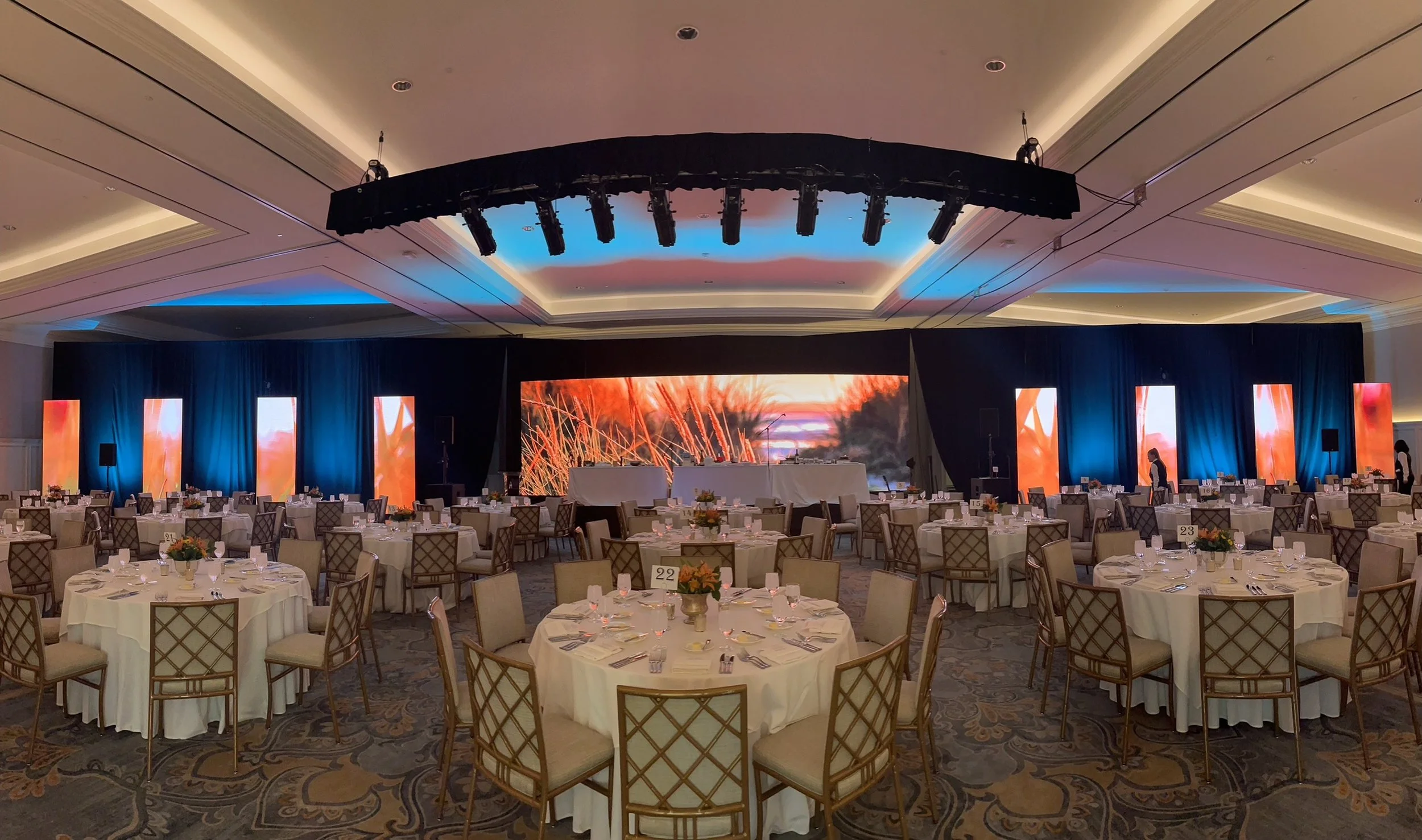 Banquet hall set up with round tables, white tablecloths, floral centerpieces, and a large stage with a digital screen displaying a sunset landscape, surrounded by black drapes and blue lighting.