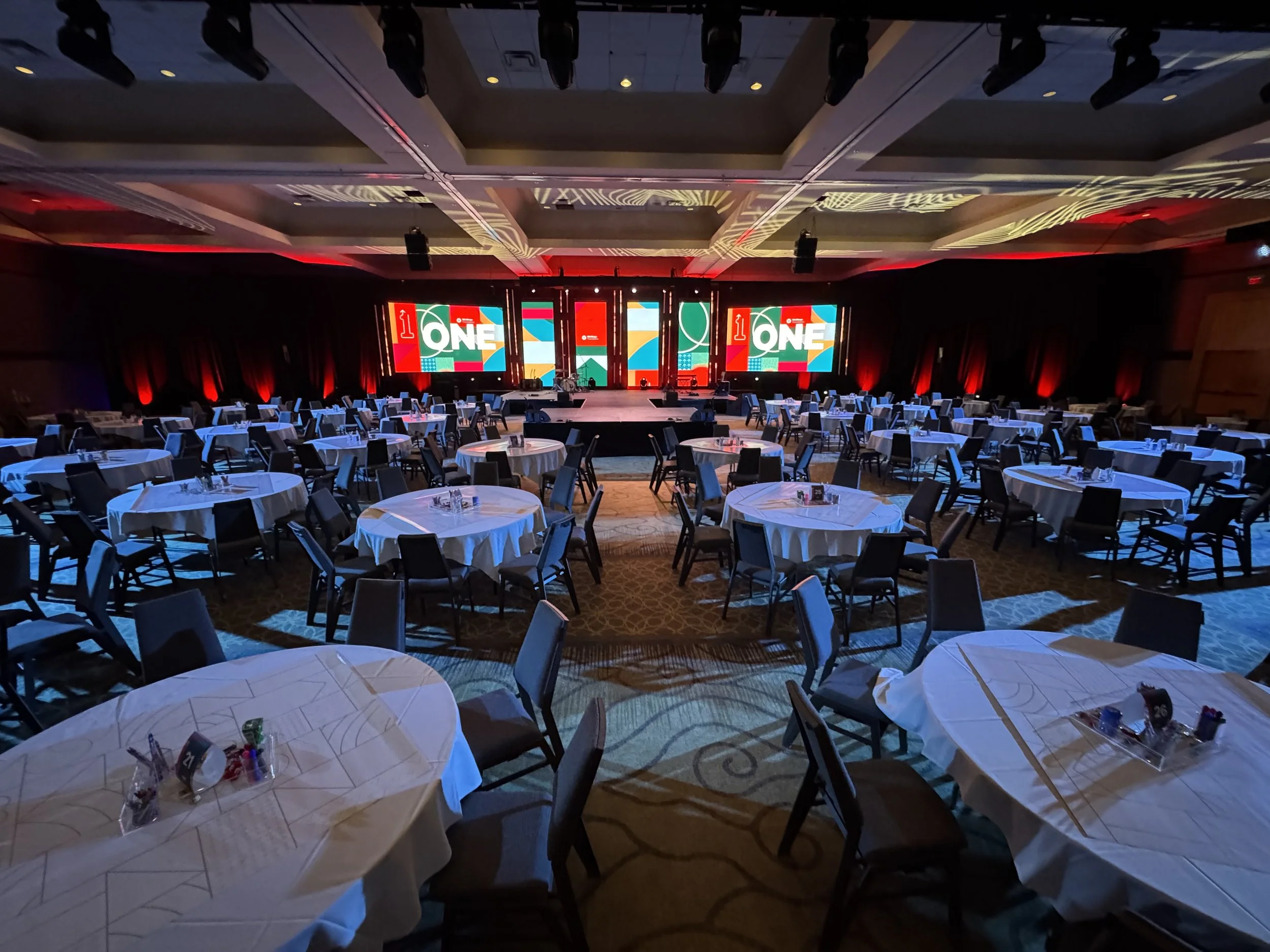 Empty banquet hall with round tables covered with white tablecloths, set up for an event, facing a stage with three large digital screens displaying colorful graphics and the word 'ONE' in the center.