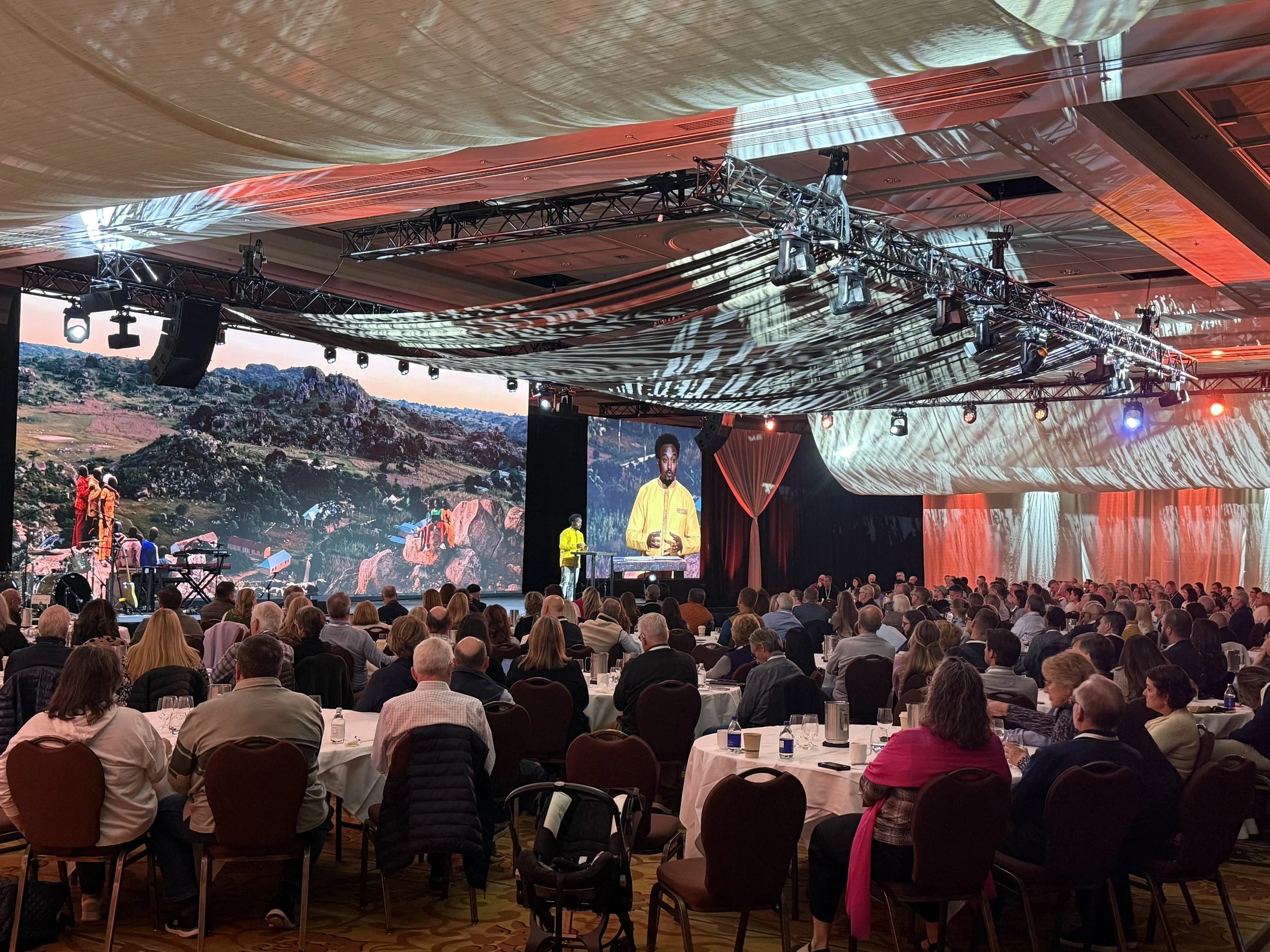 An indoor conference with a large seated audience watching a stage presentation, large screens, and scenic background images.