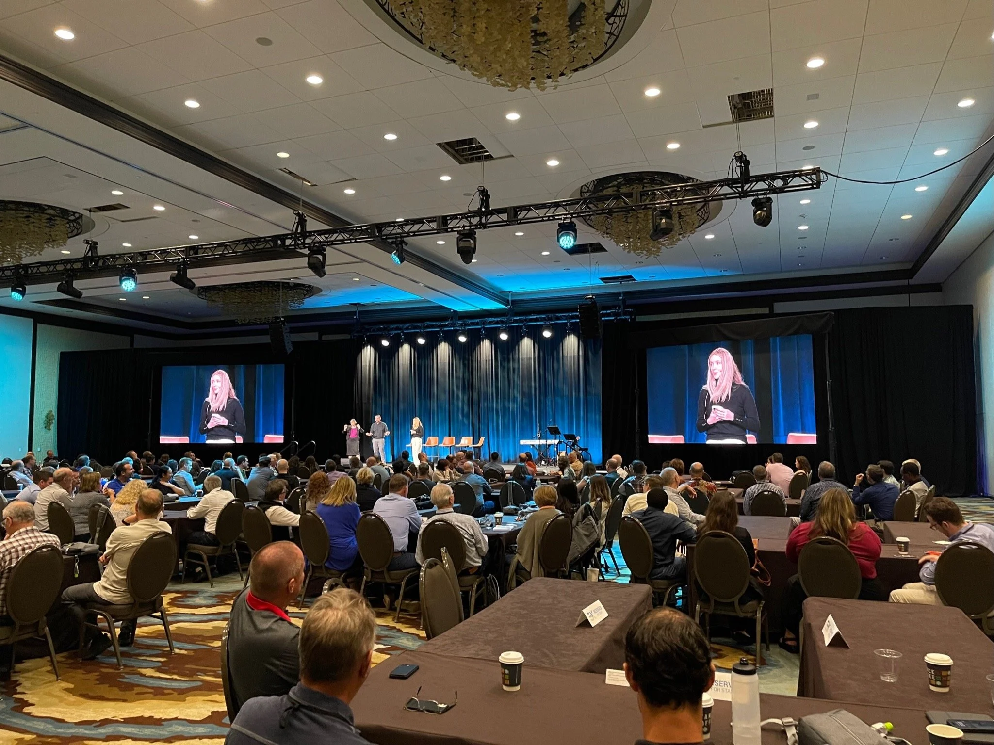 A large conference room filled with attendees watching a presentation on stage. There are two large screens showing a woman speaking, with a blue curtain backdrop behind her. The ceiling has stage lights and decorations, and there are tables with coffee cups and personal items in the foreground.