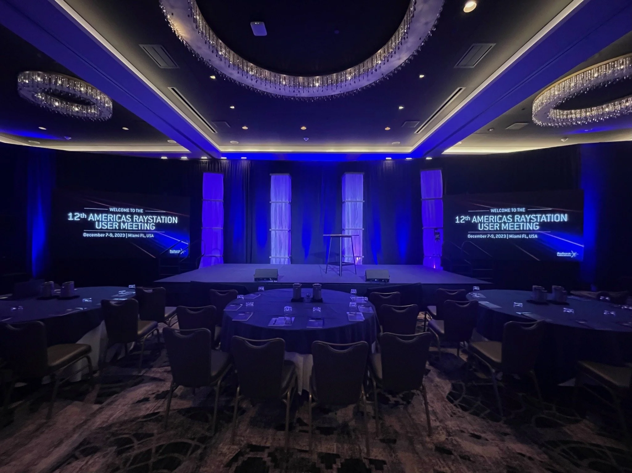Conference room set up for the 12th Americas Raystation User Meeting, with a stage, large screens displaying event information, and tables with chairs arranged for attendees.