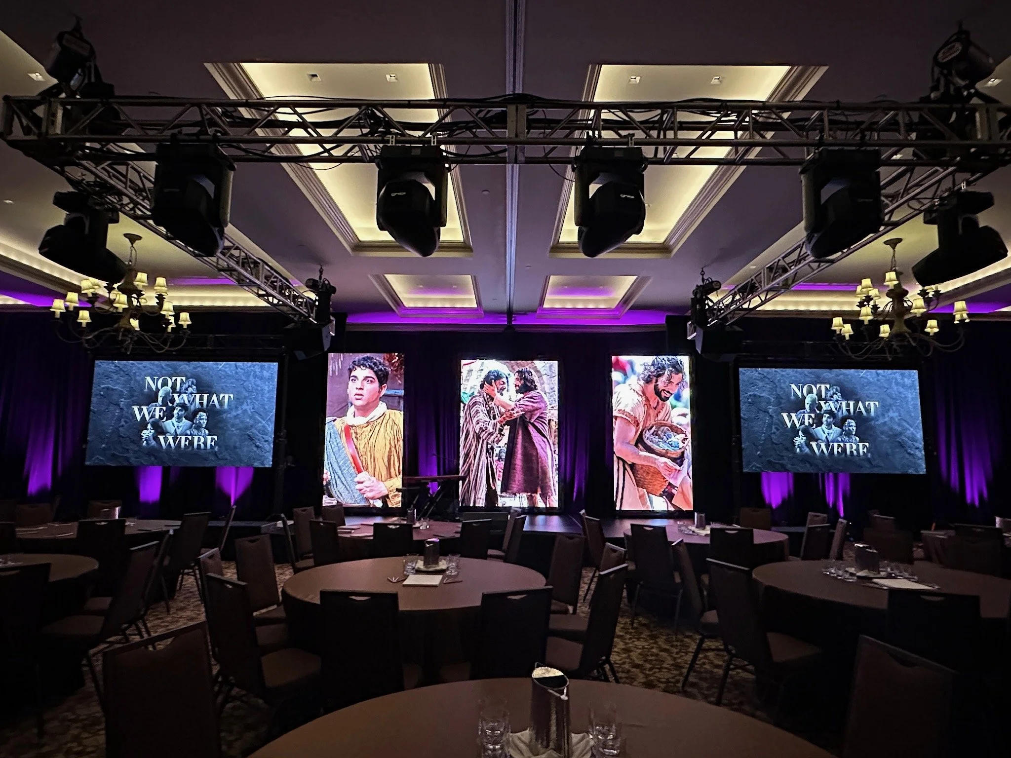 Empty banquet hall with round tables set for an event, large screens and stage backdrop with theatrical posters and the text 'Not What We Were' in a dimly lit room with purple lighting.