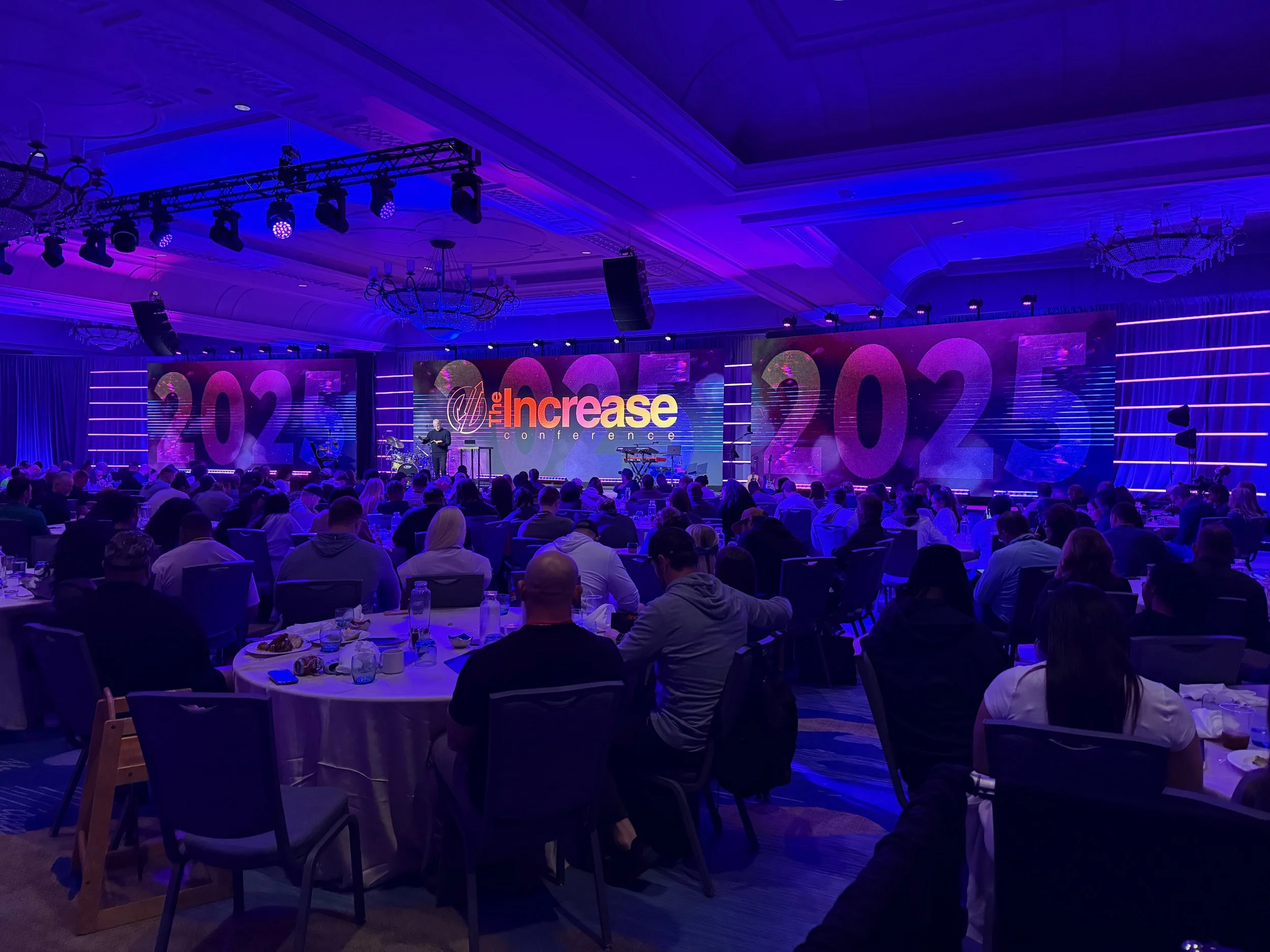 Conference hall with audience watching a speaker on stage, displaying the text 'The Increase Conference 2025' on large screens.