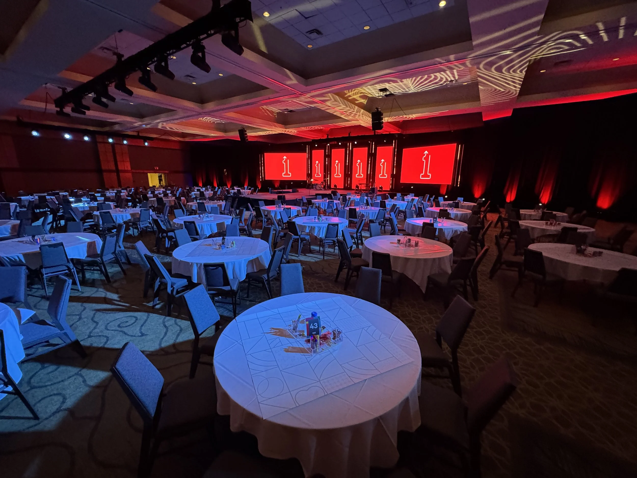 An empty banquet hall with round tables covered in white tablecloths, set up for an event with a stage at the front displaying multiple large red screens, illuminated with red and blue lighting.