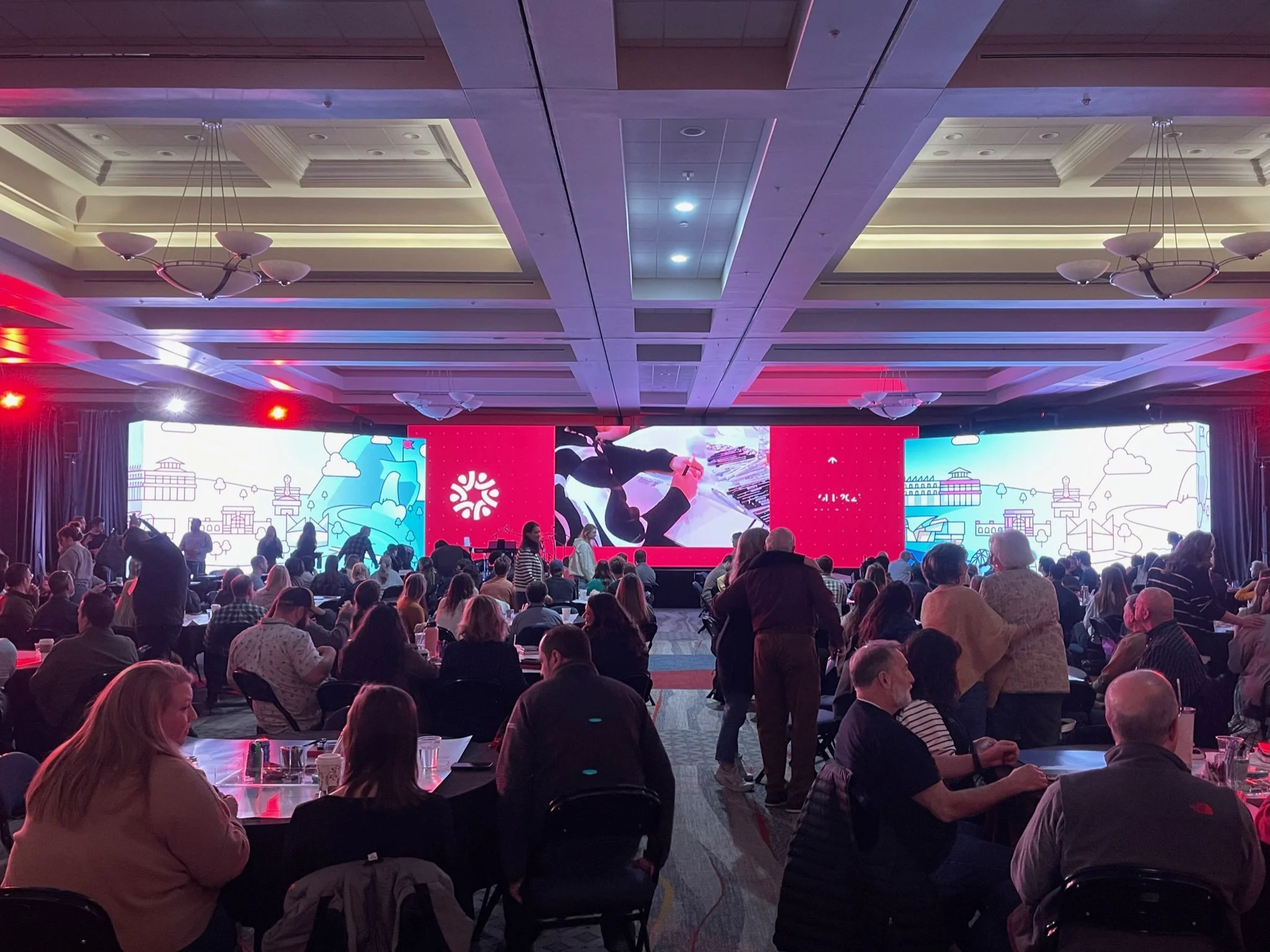 Conference room with many people seated at tables, facing a stage with large screens displaying graphics and a red logo, with some people standing and talking.