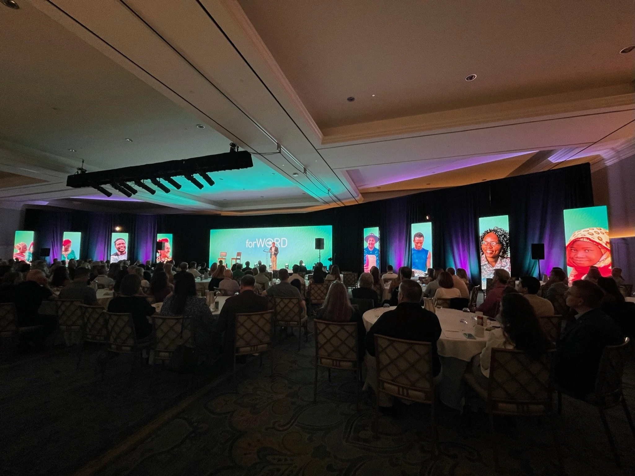 A large conference or event with an audience seated at round tables in a dimly lit ballroom. A stage at the front features a speaker talking, with multiple large screens displaying various images of diverse people.
