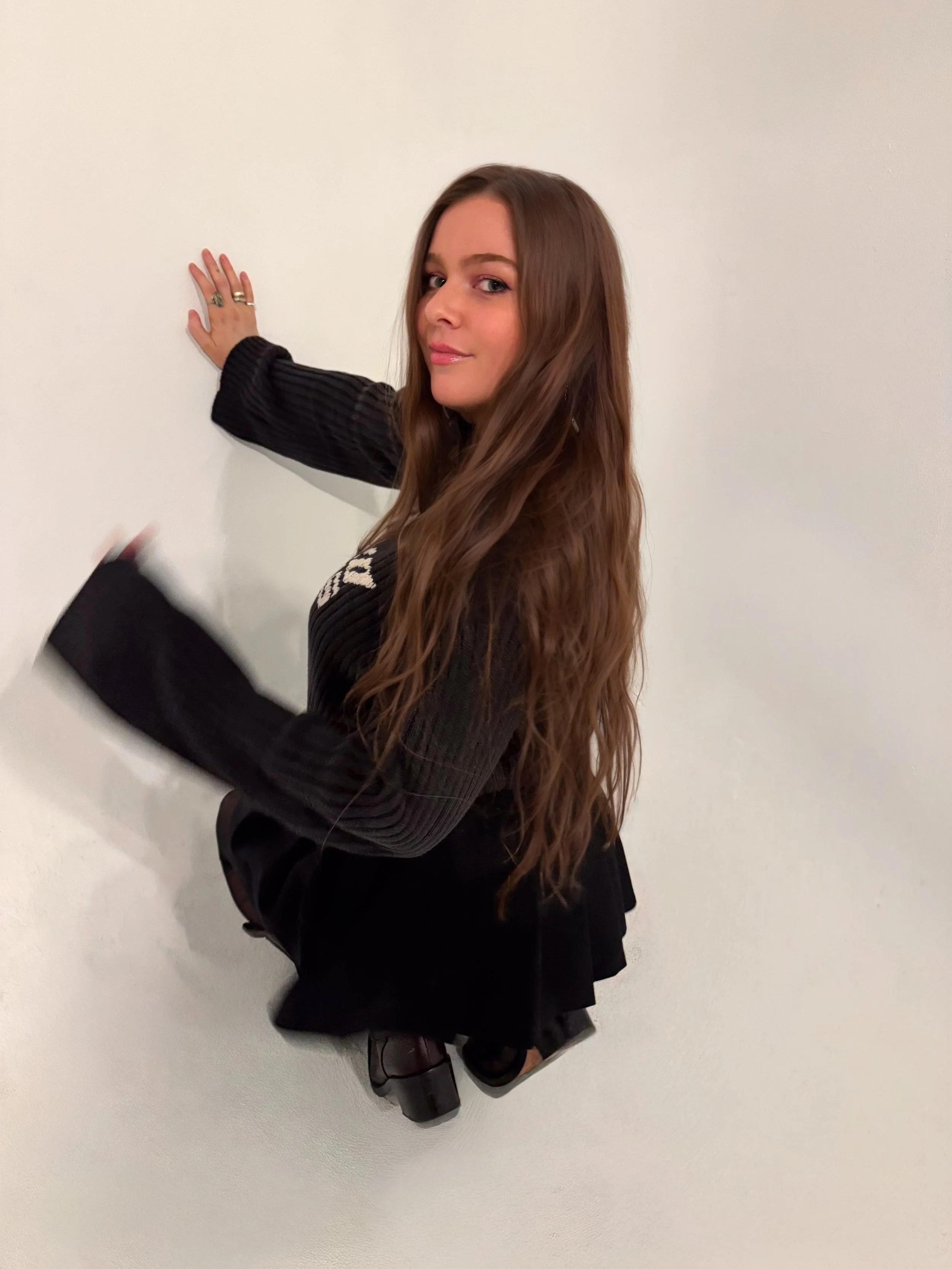 Woman in a black outfit posing against a white wall with one hand raised.