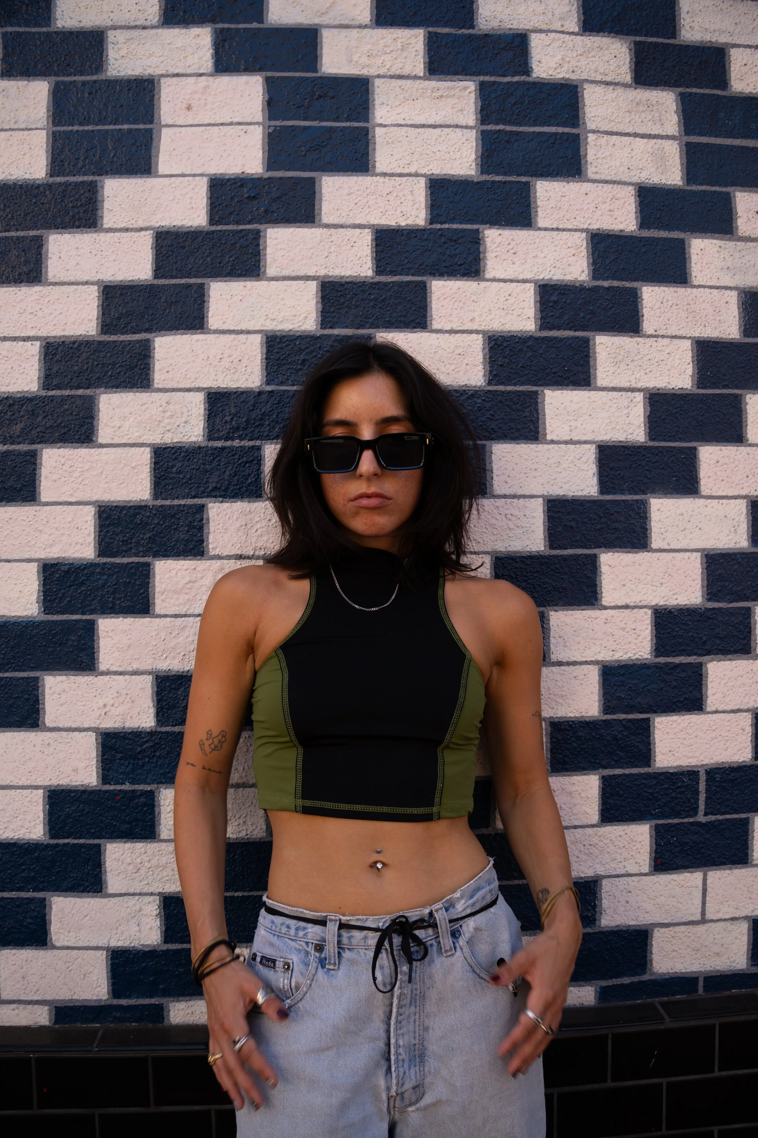 Person wearing sunglasses, a black-and-olive crop top, and light jeans standing against a blue-and-white brick wall