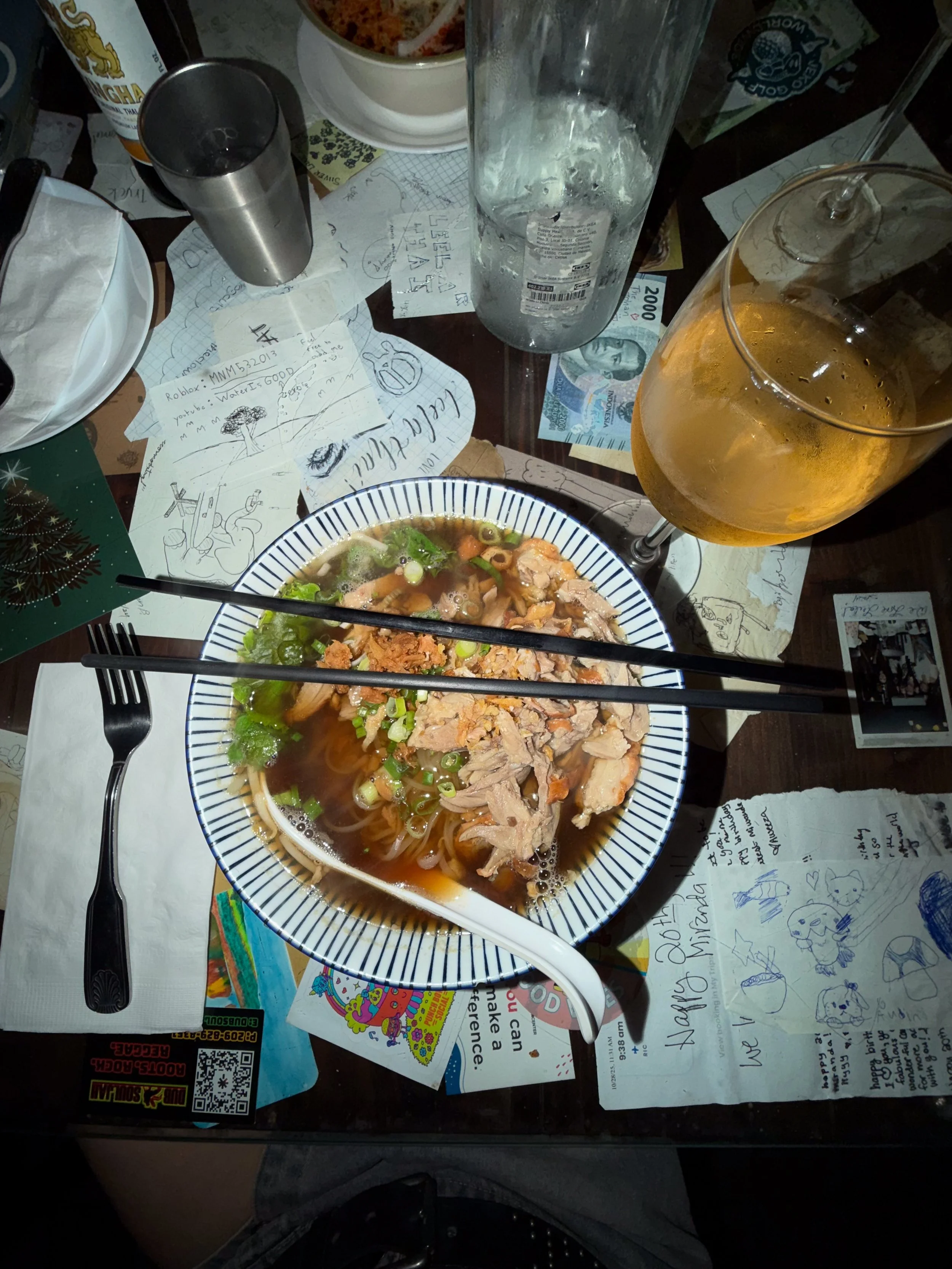 Bowl of Vietnamese noodle soup with sliced meat and herbs, chopsticks, orange drink, and condiments on a cluttered dining table