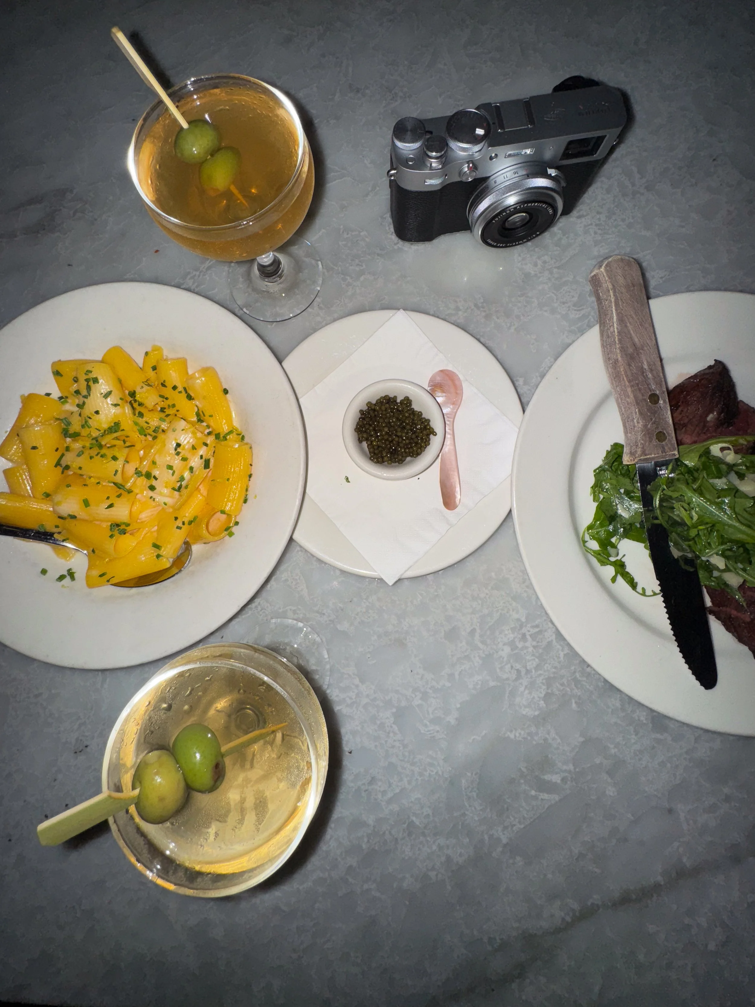 Overhead view of a dining table with cocktails garnished with green olives, pasta, caviar, salad, steak, and a vintage camera.