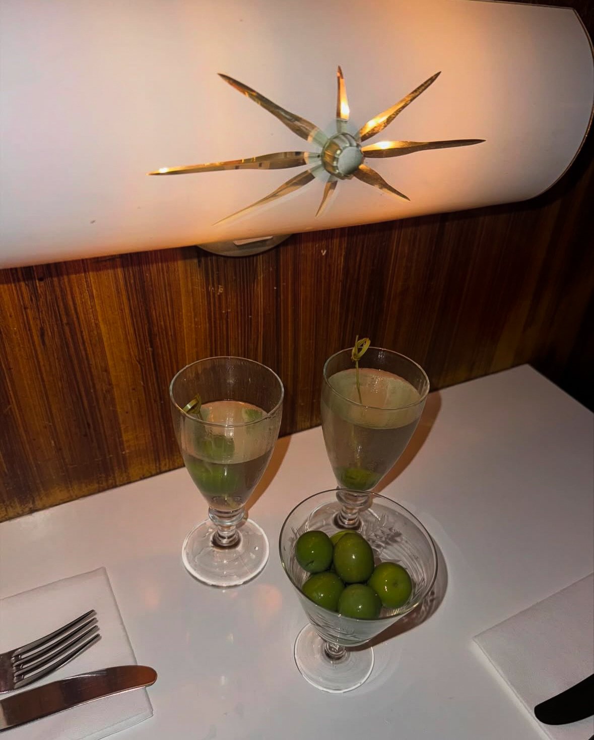 Two cocktails garnished with lime sit beside a bowl of green olives on a restaurant table beneath a starburst wall light.