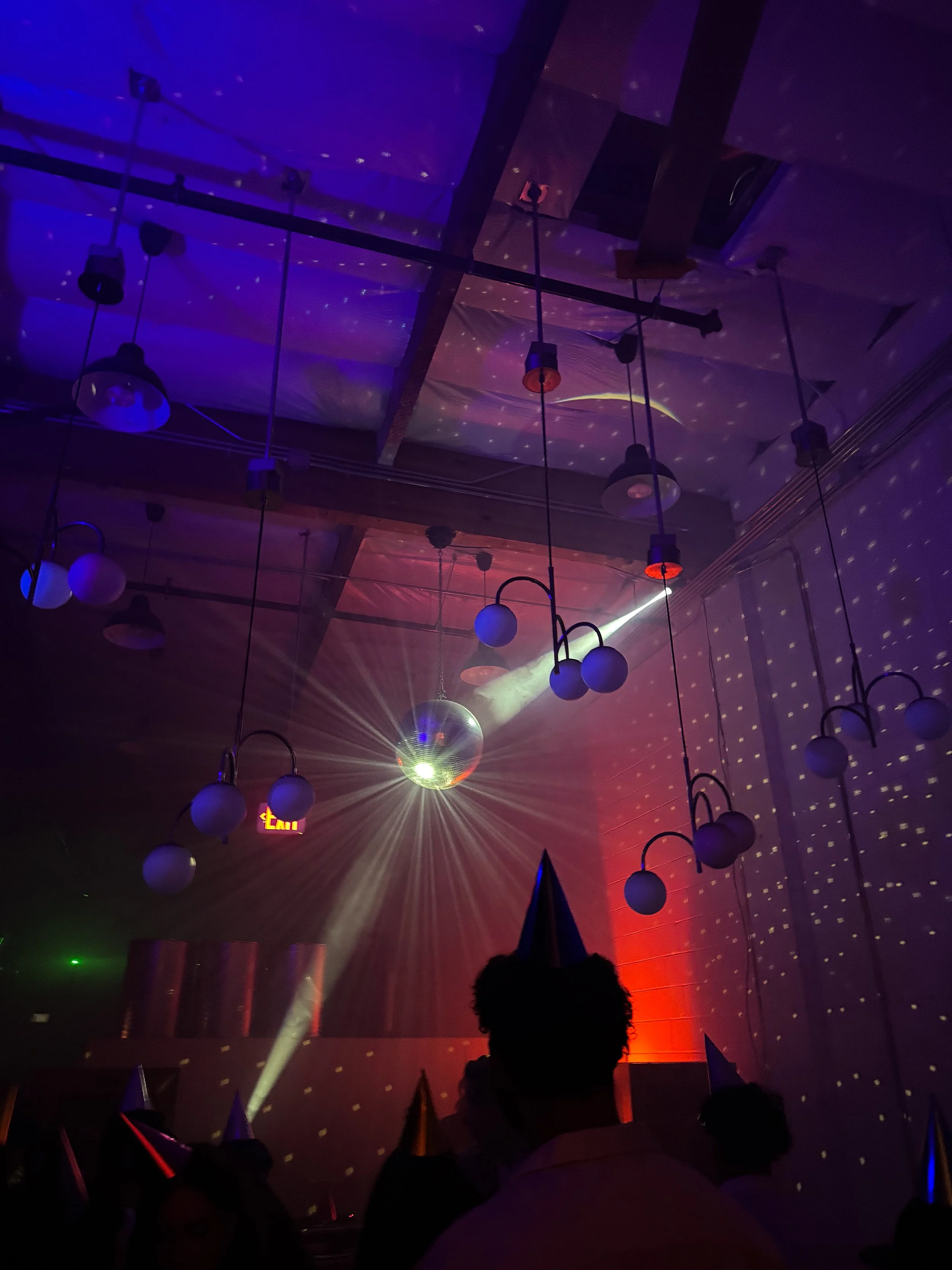 Darkly lit party venue with hanging spherical lights, disco ball, star projections, colorful beams, and silhouetted guests wearing cone party hats