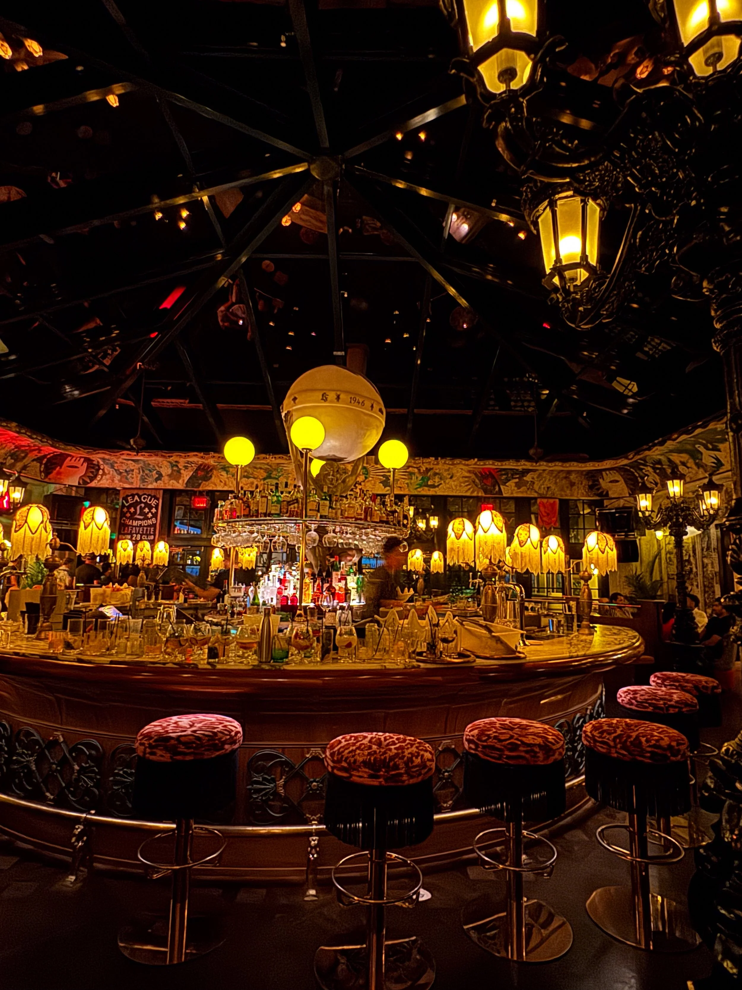 Dimly lit, ornate bar interior with glowing lamps, a mirrored back bar, colorful wall murals, and leopard-print bar stools.
