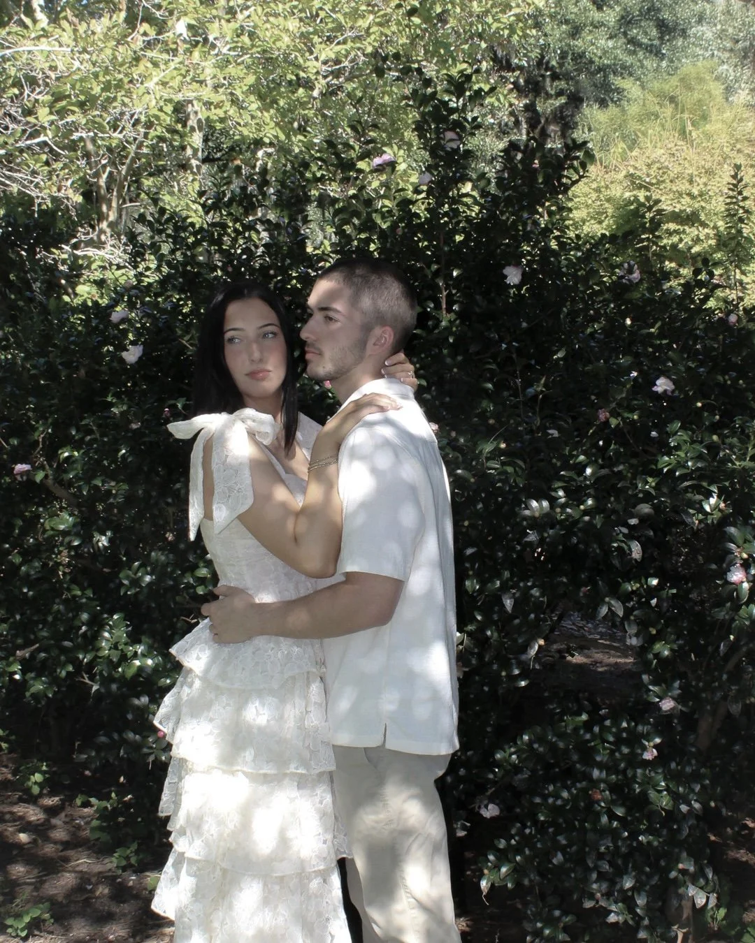 A couple in white clothing, standing close together among green bushes and trees with sunlight filtering through, creating a serene outdoor setting.