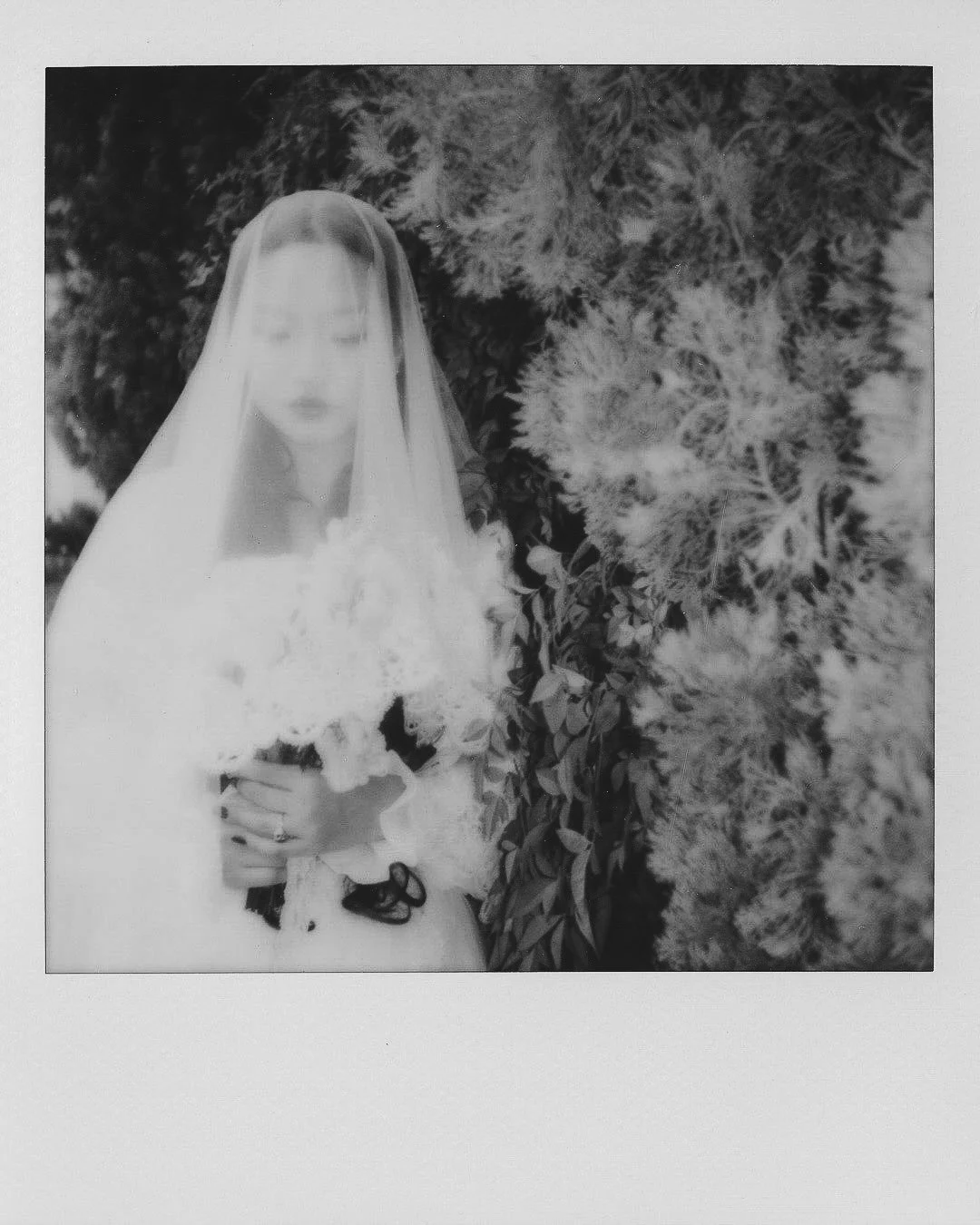 Polaroid of a bride with long hair in a garden in black and white. Her veil covers her face and she holds a bouquet.