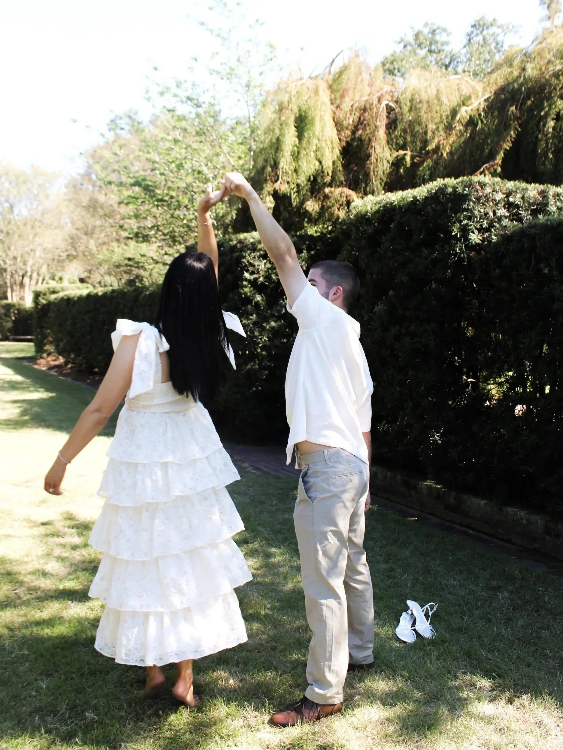 Woman and man dancing in garden, both wearing white outfits. Greenery surrounds them.