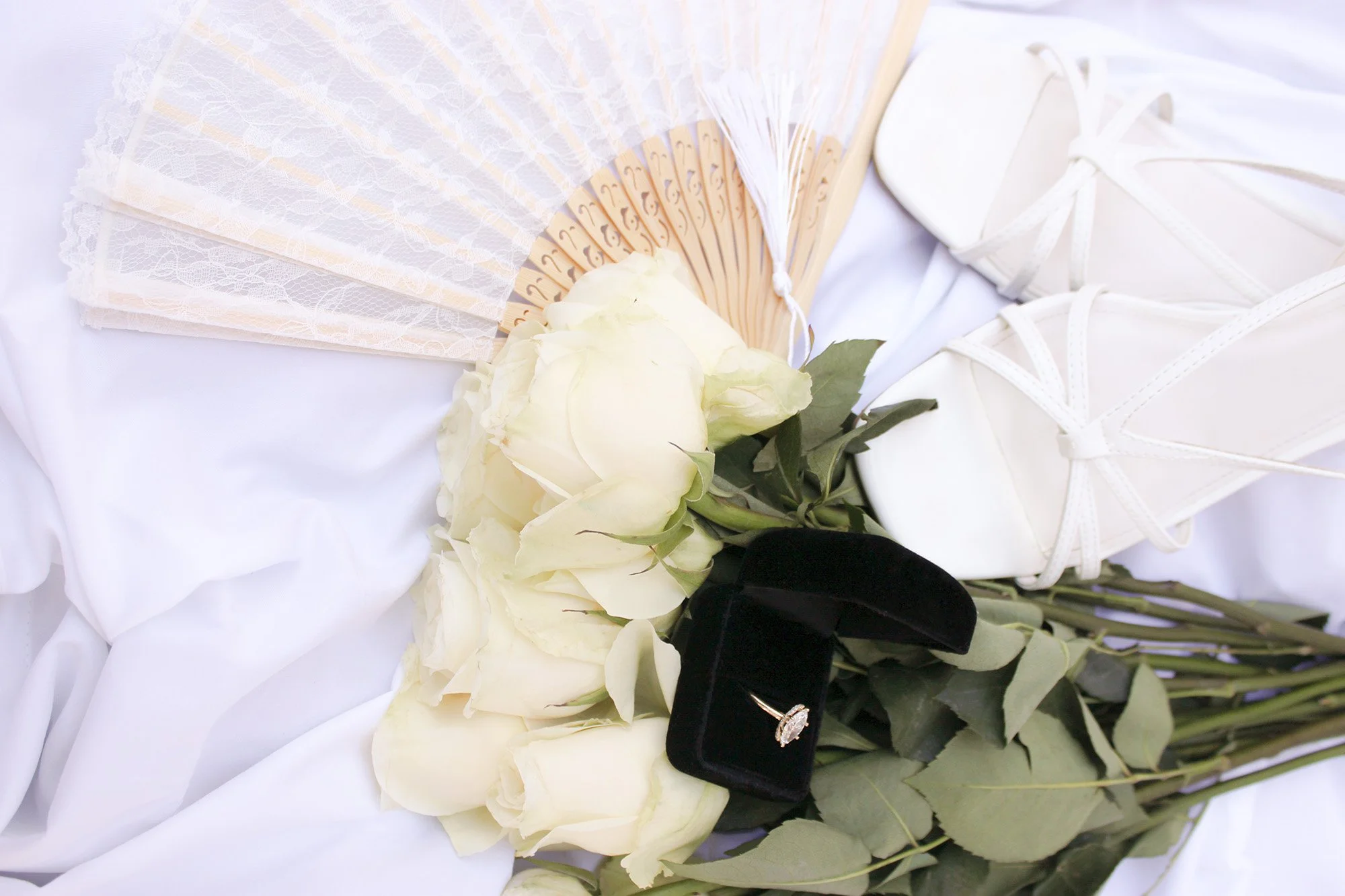 Close up of a white wedding bouquet with white roses, a black ring box with an engagement ring, a lace fan, and a pair of white bridal shoes on a white fabric background.