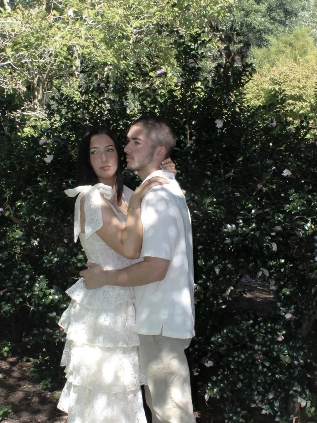 Woman and man wearing white hugging each other and looking into the distance standing in garden.